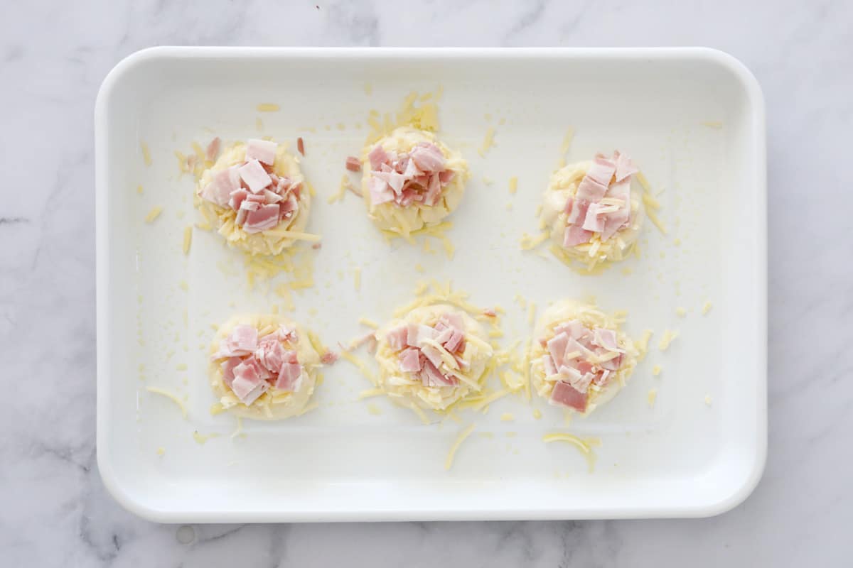 Rolls on a baking tray topped with grated cheese and chopped bacon.