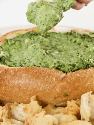 A hand lifting up a piece of creamy spinach cob loaf dip with crusty bread.