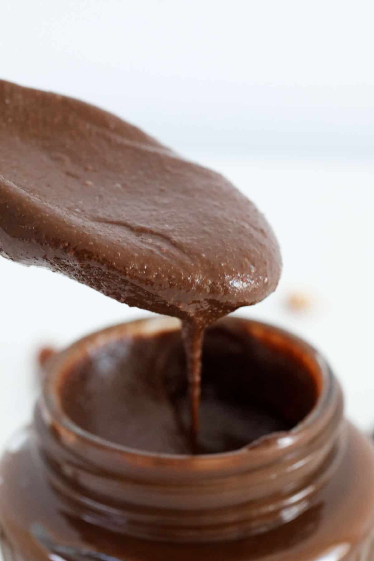 Smooth homemade Nutella dripping from a spoon held above a jar.