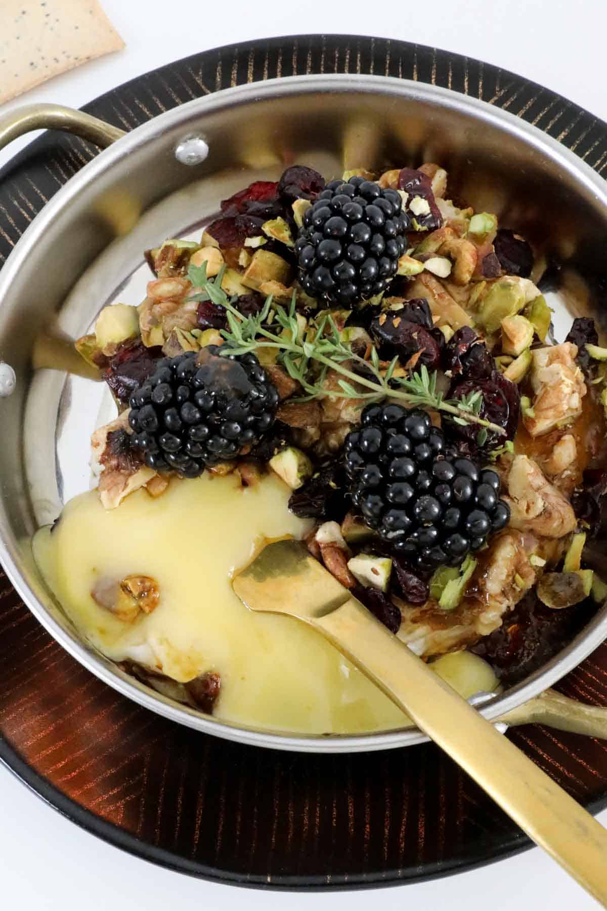 A melted baked cheese in a small round baking dish topped with nuts and fresh blackberries.