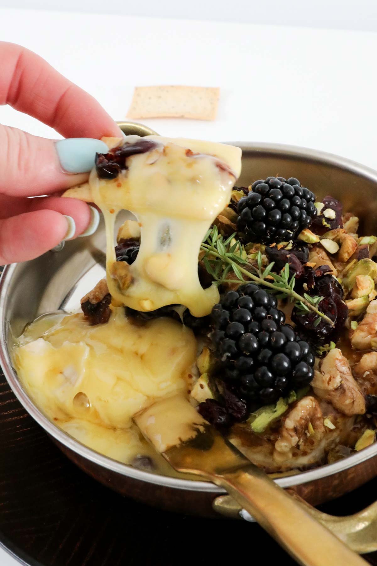 A hand holding a cracker dipped in melted cheese with blackberries.