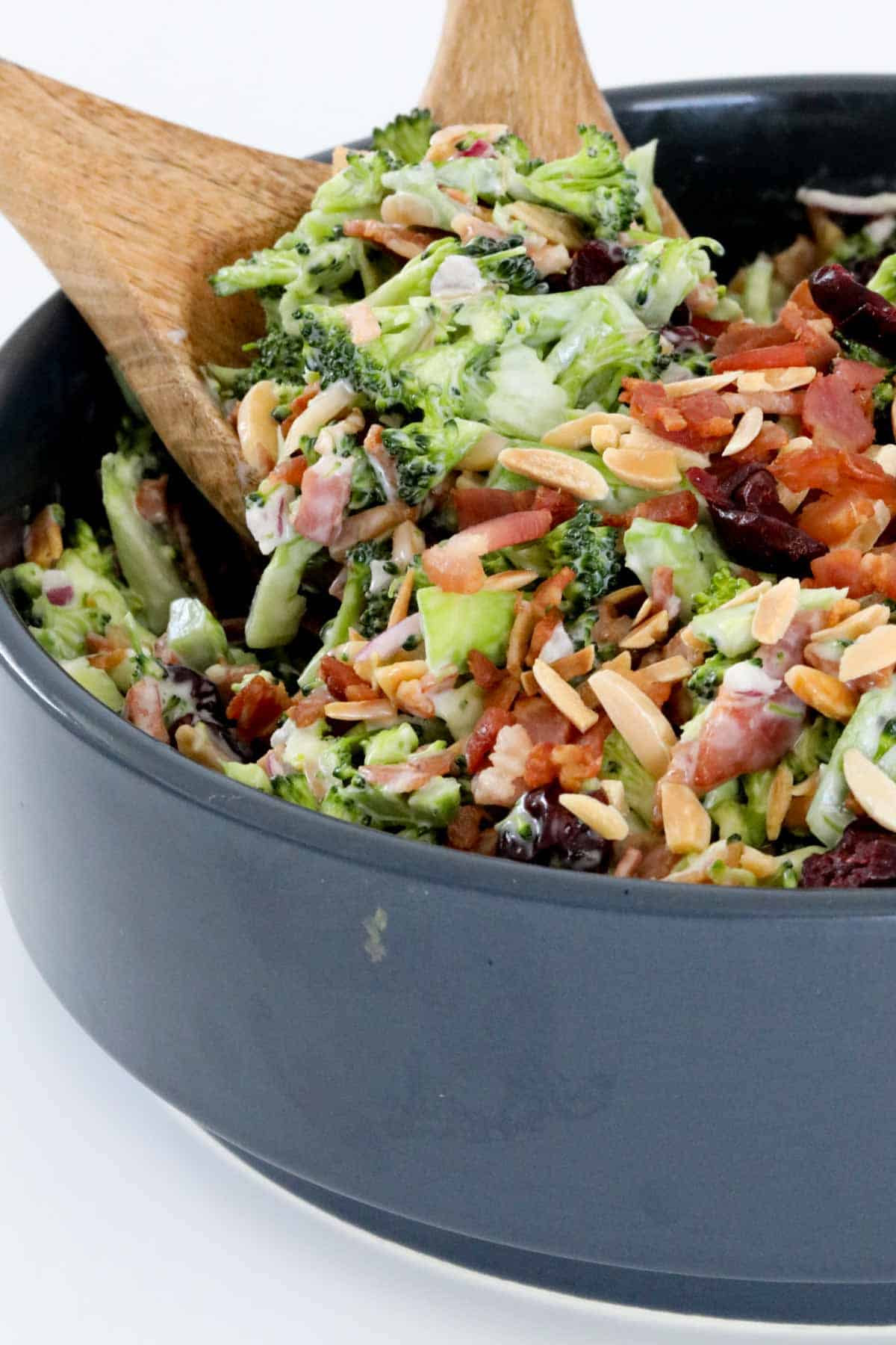 Two wooden salad servers in a bowl of crunchy broccoli salad with slivered almonds and cranberries.