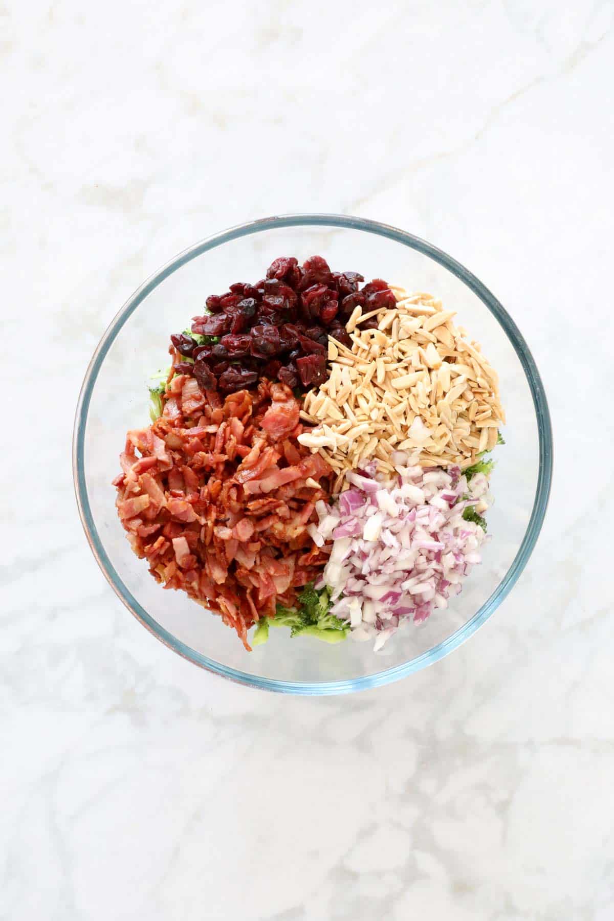 Crispy bacon, broccoli, dried cranberries, slivered almonds and diced red onions in a glass bowl.
