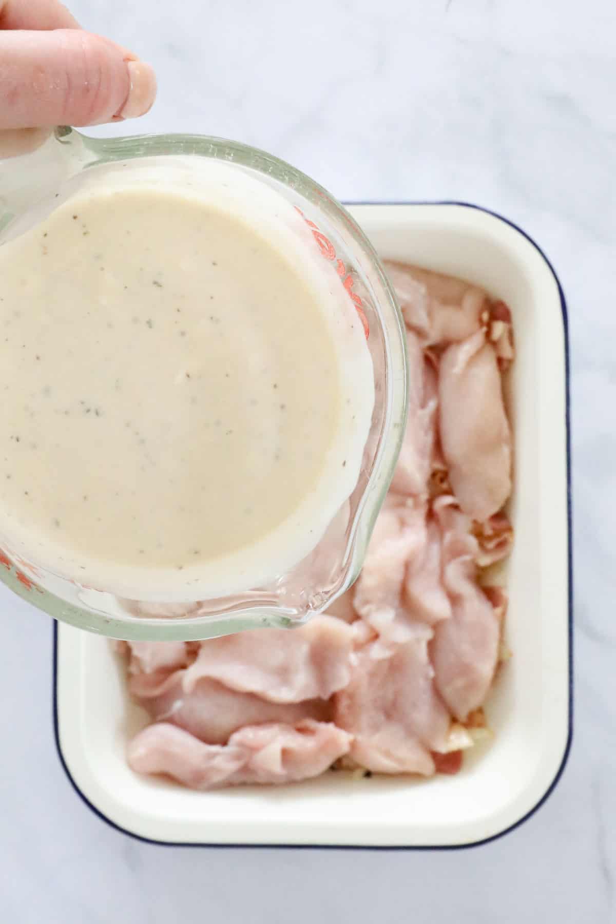 A hand pouring a glass of creamy ranch dressing and sour cream sauce over chicken in a baking dish. 