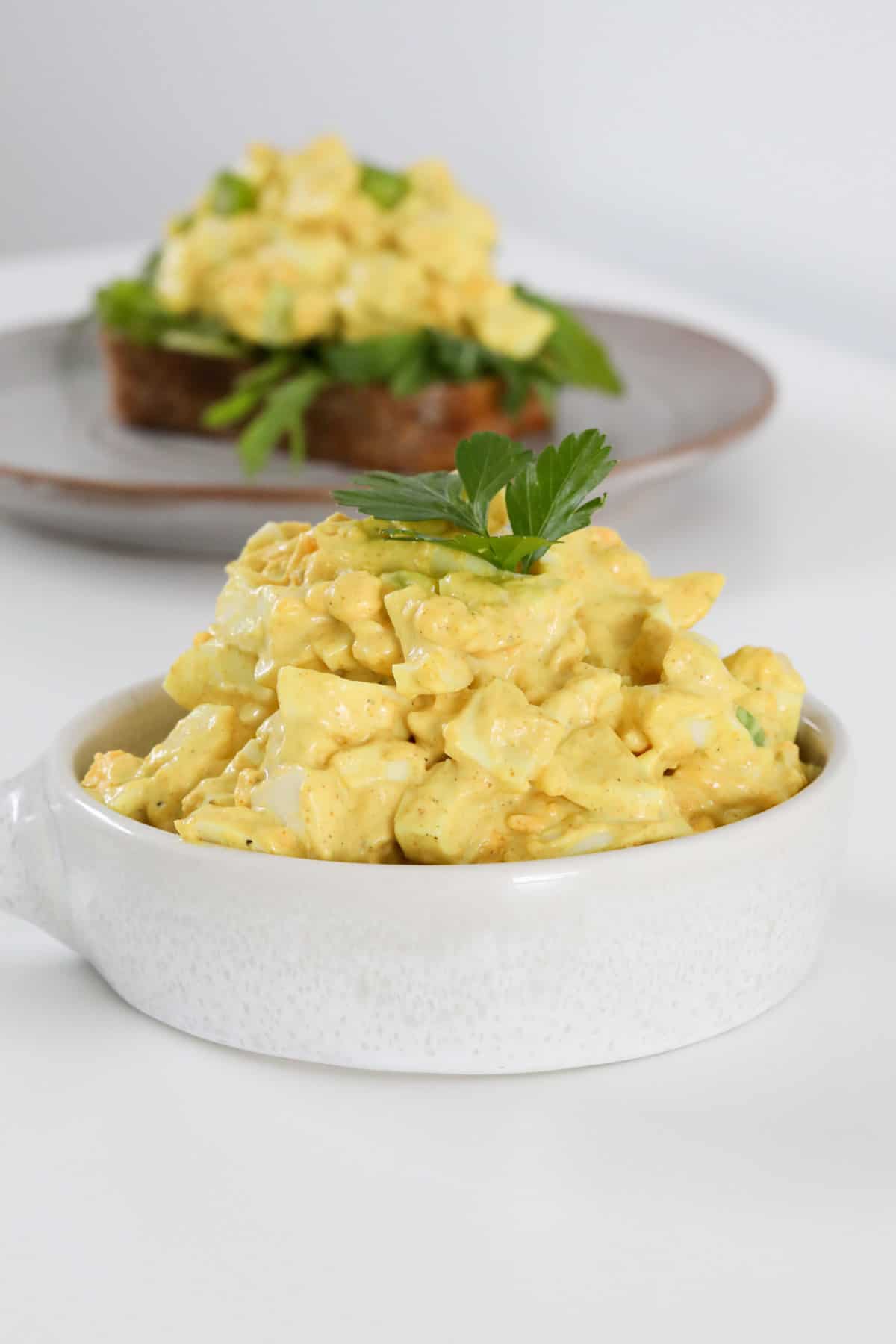 Curriend egg salad in a white bowl with a sprig of parsley on top.