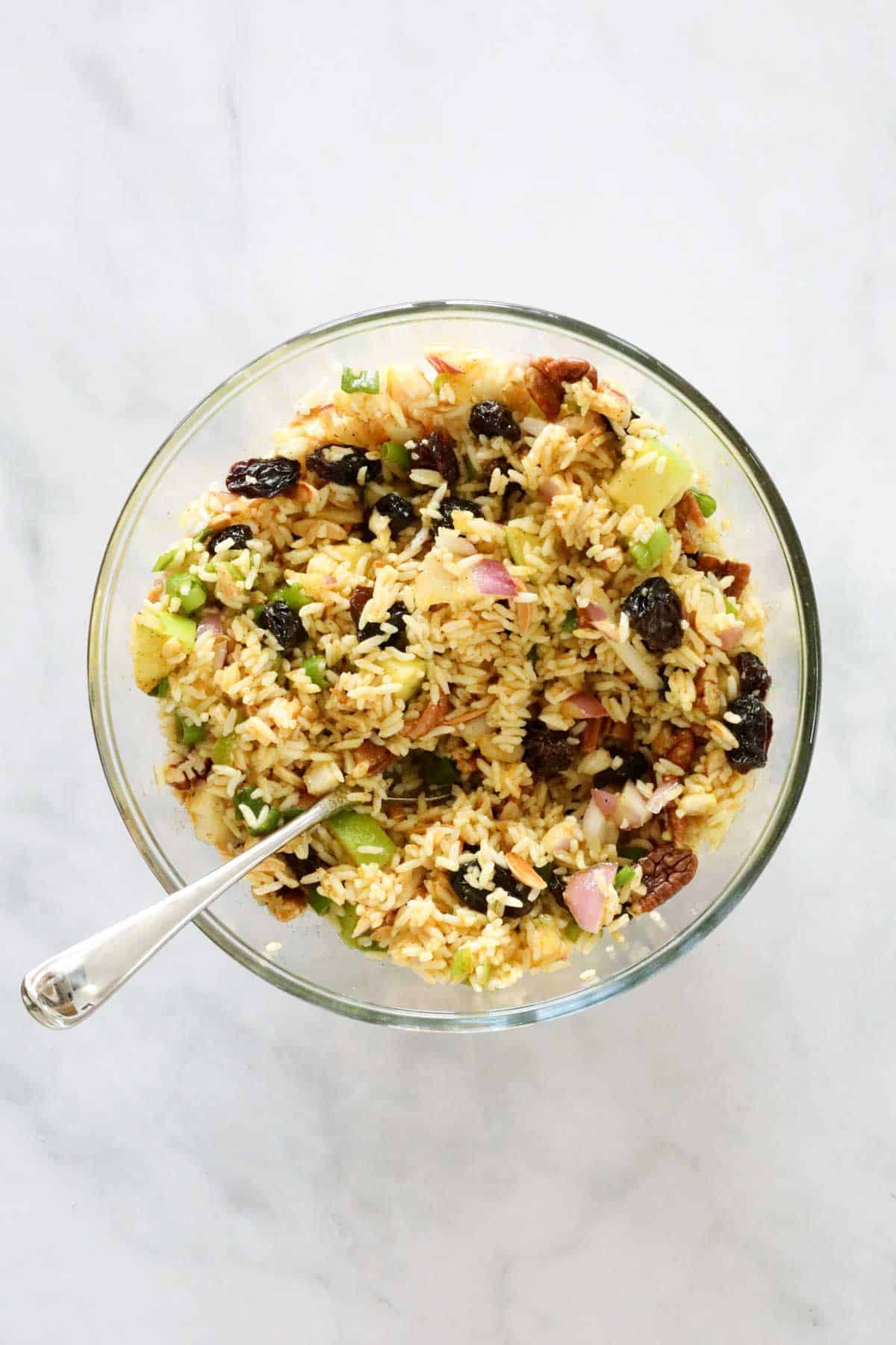 A large glass bowl filled with rice, spring onions, raisins, toasted nuts and mixed together with dressing.
