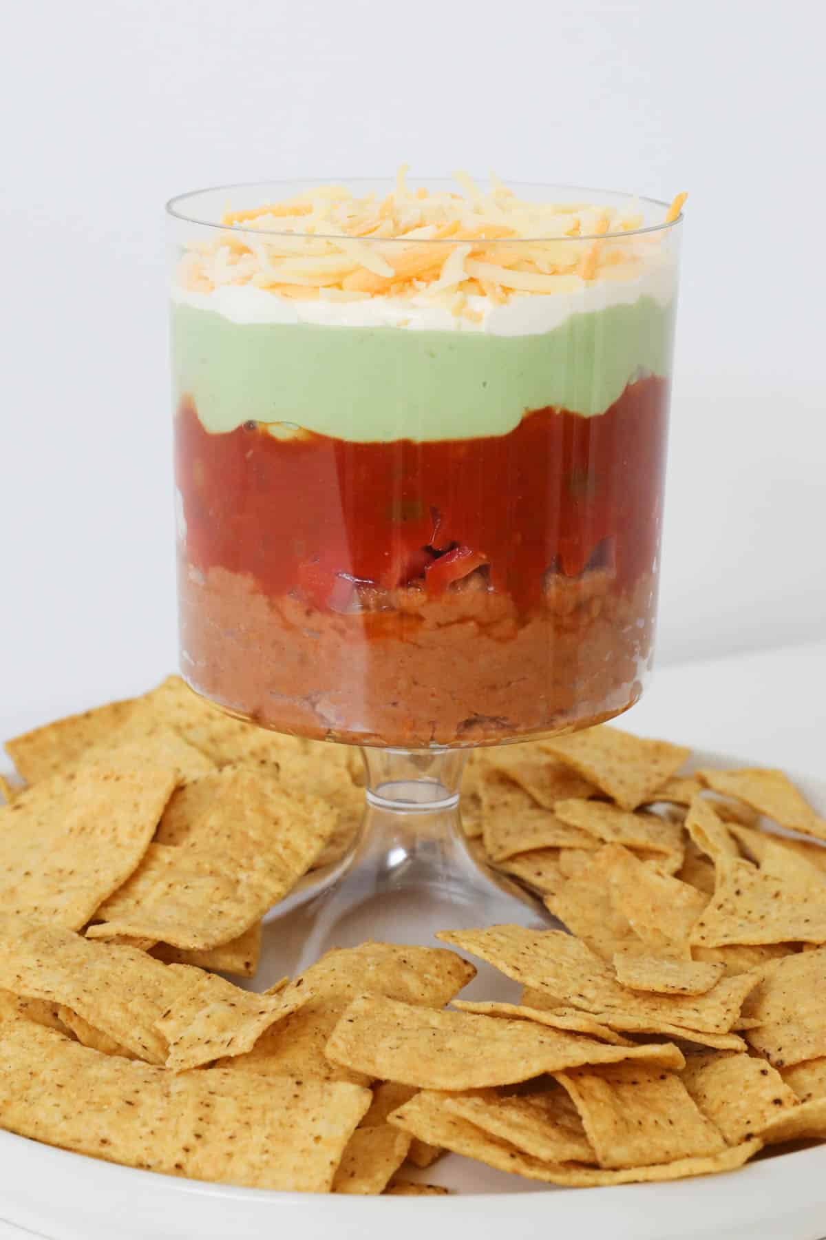 A tall bowl holding a colouful Mexican layer dip surrounded by tortilla strips.