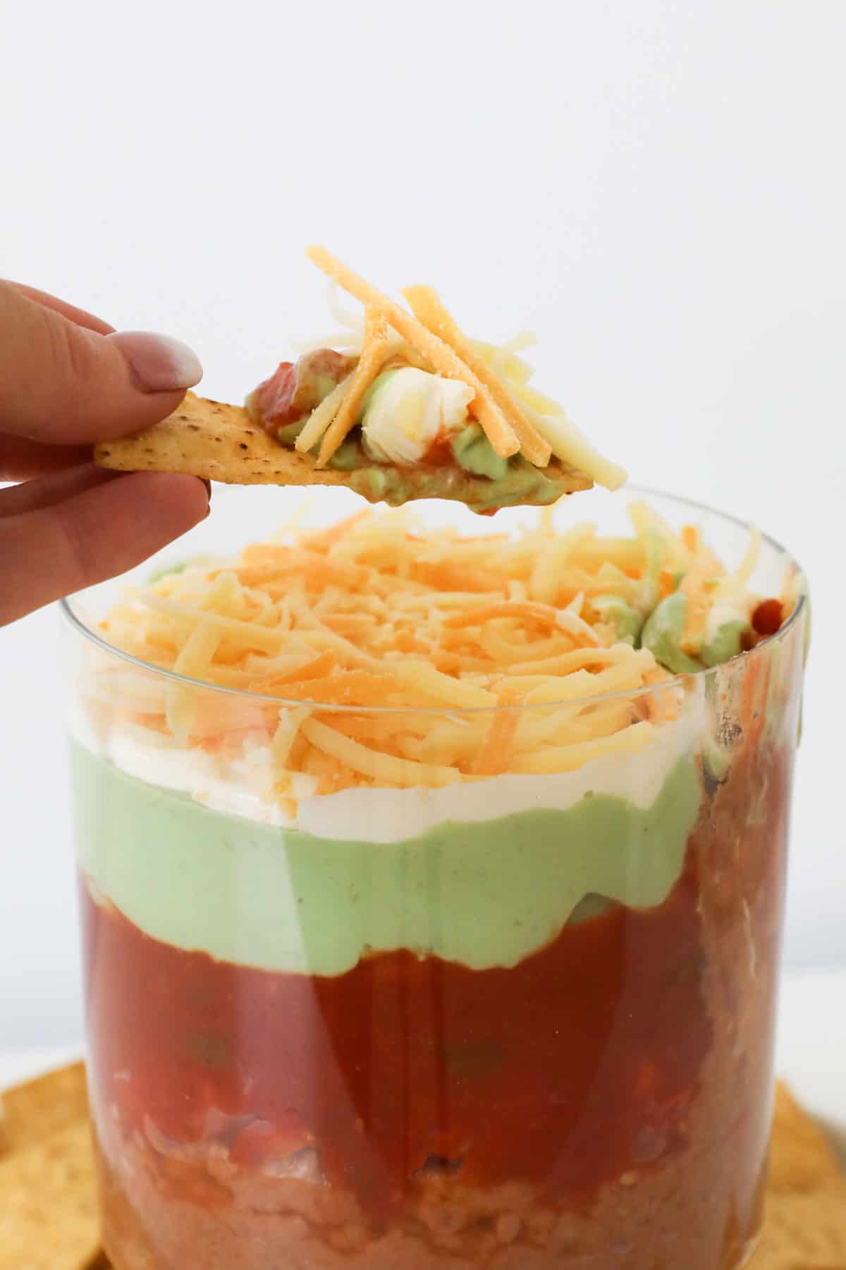 A hand holding a corn chip with dip over a clear bowl of colorful mexican layer dip..