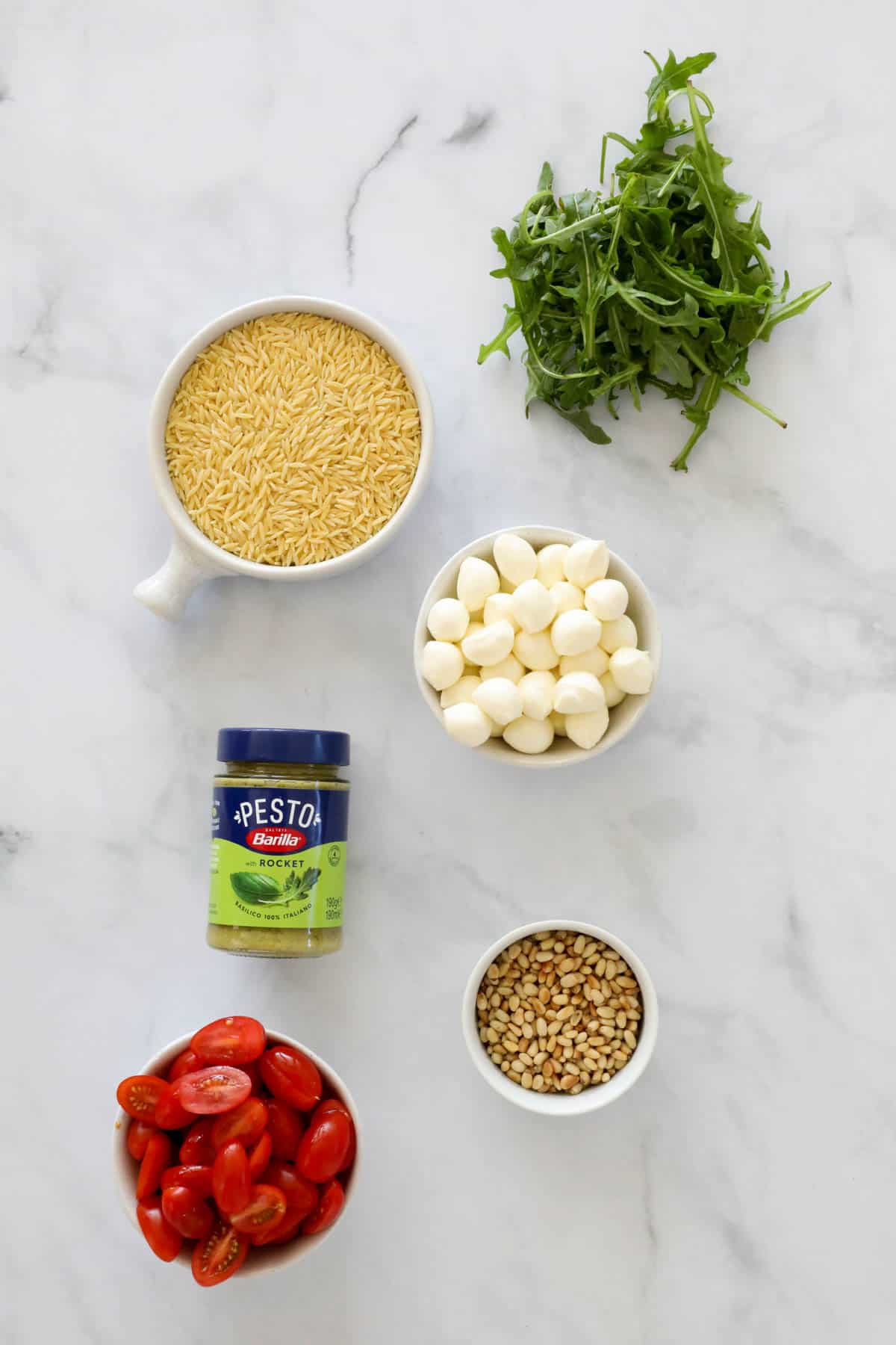 Ingredients to make a pasta pesto salad in small bowls on a bench top.