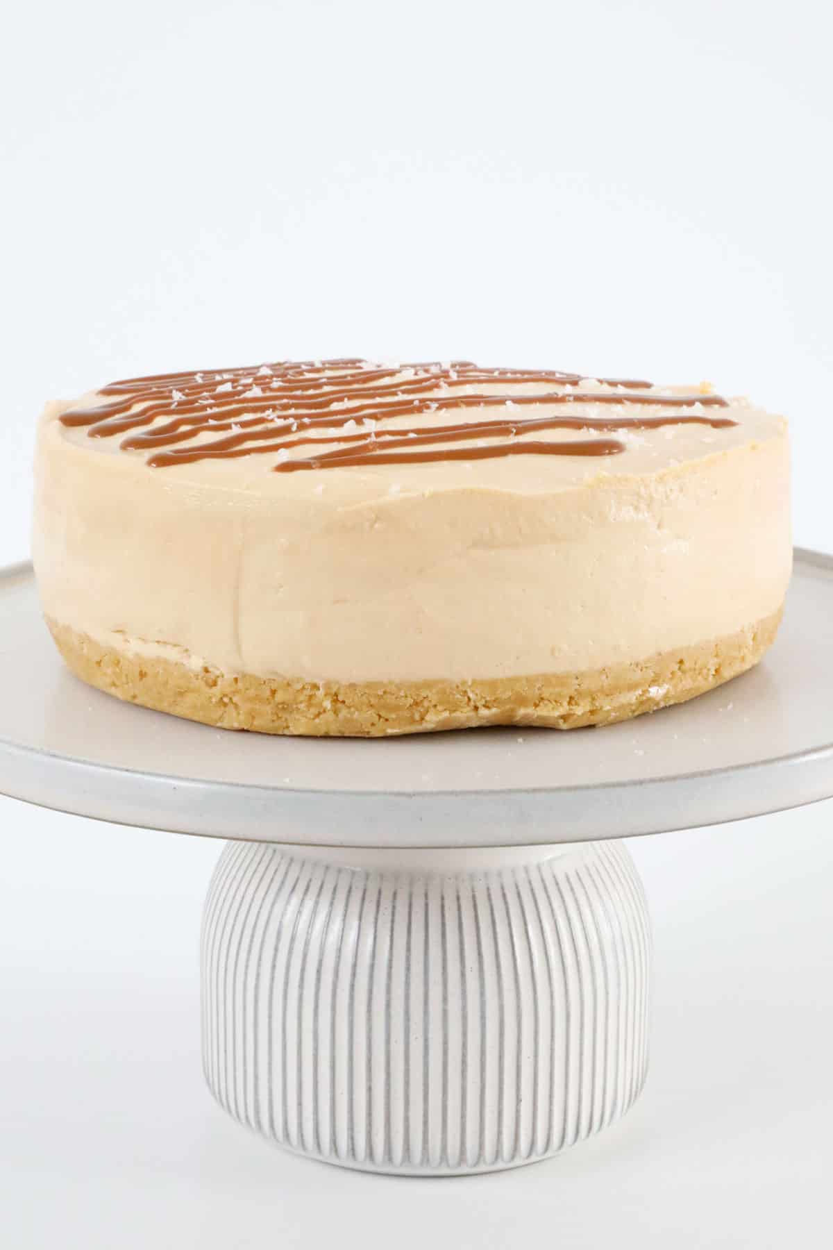 A salted caramel cheesecake, drizzled with caramel sauce, on a cake stand.