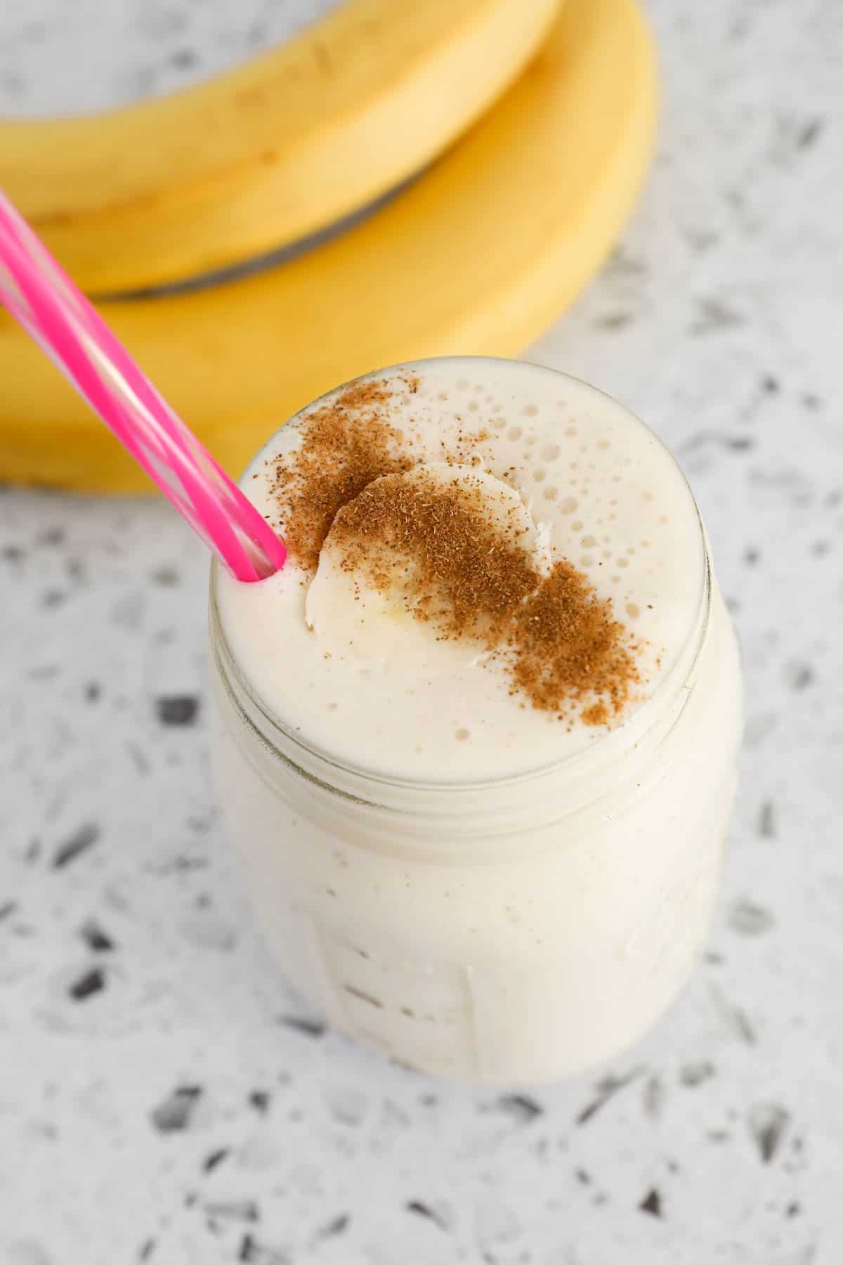 A banana smoothie in a glass jar with a straw, in front of fresh bananas.