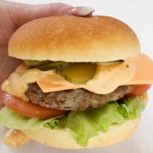 A hand holding a beef burger with cheese, tomato, lettue, pickles and sauce.
