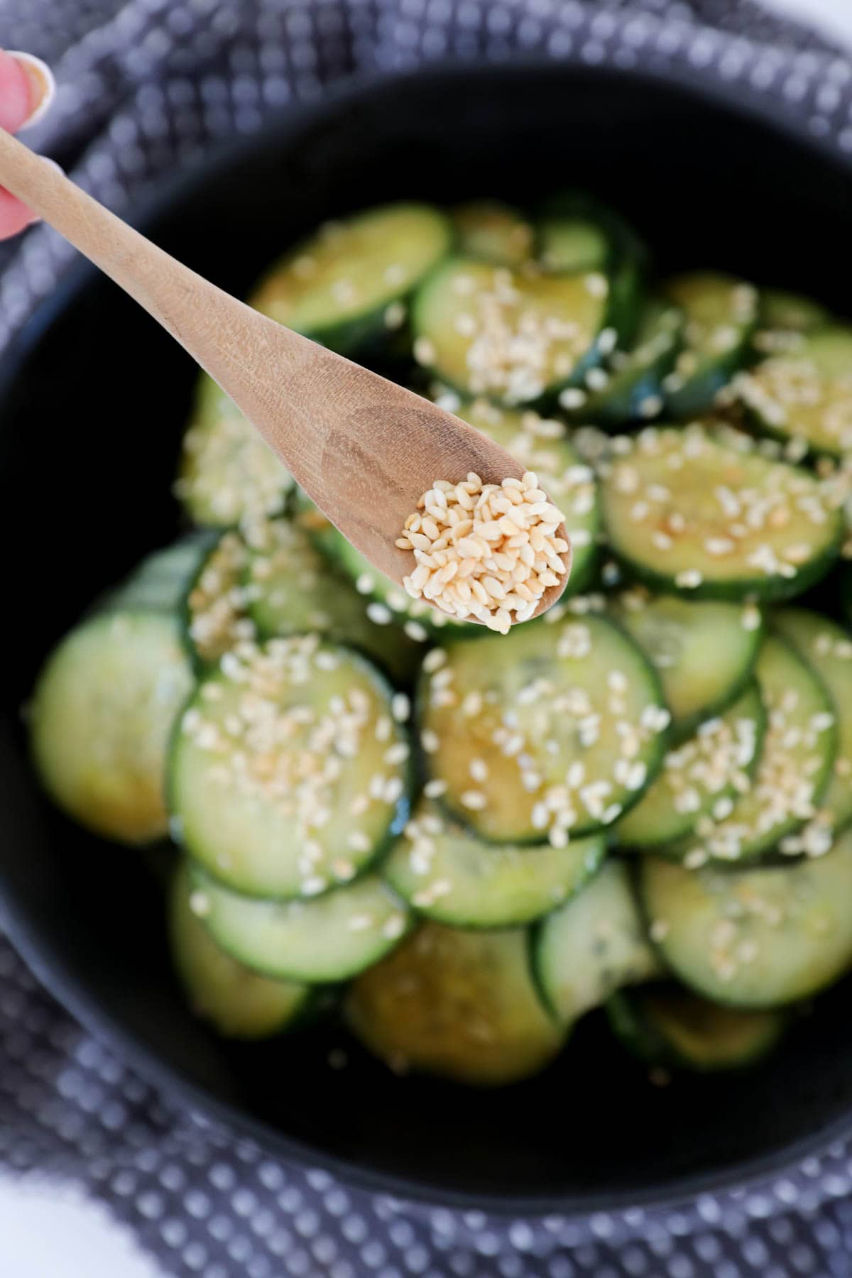 Toasted sesame seeds being sprinkled on sliced cucumbers with a wooden spoon.