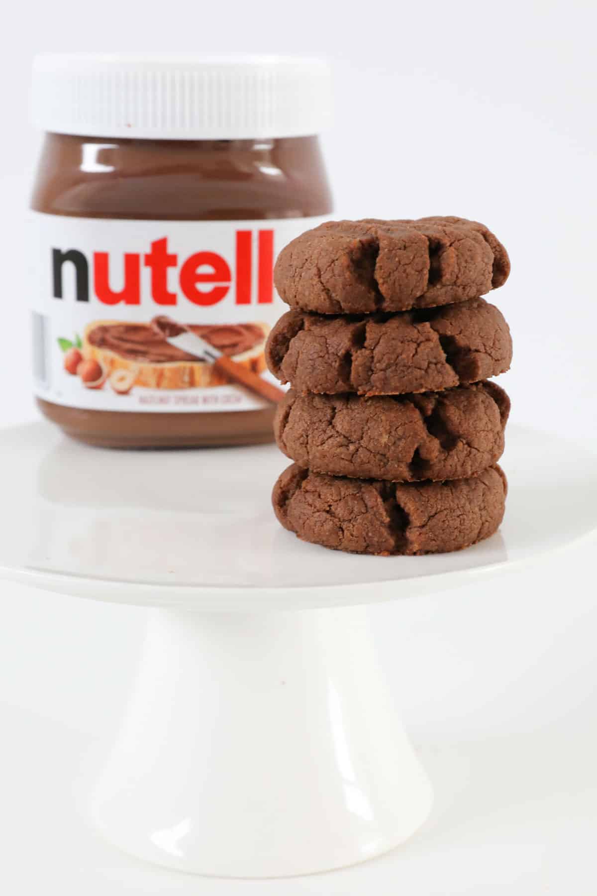 A stack of four cookies on a white cake stand with a jar of hazelnut spread in the background.