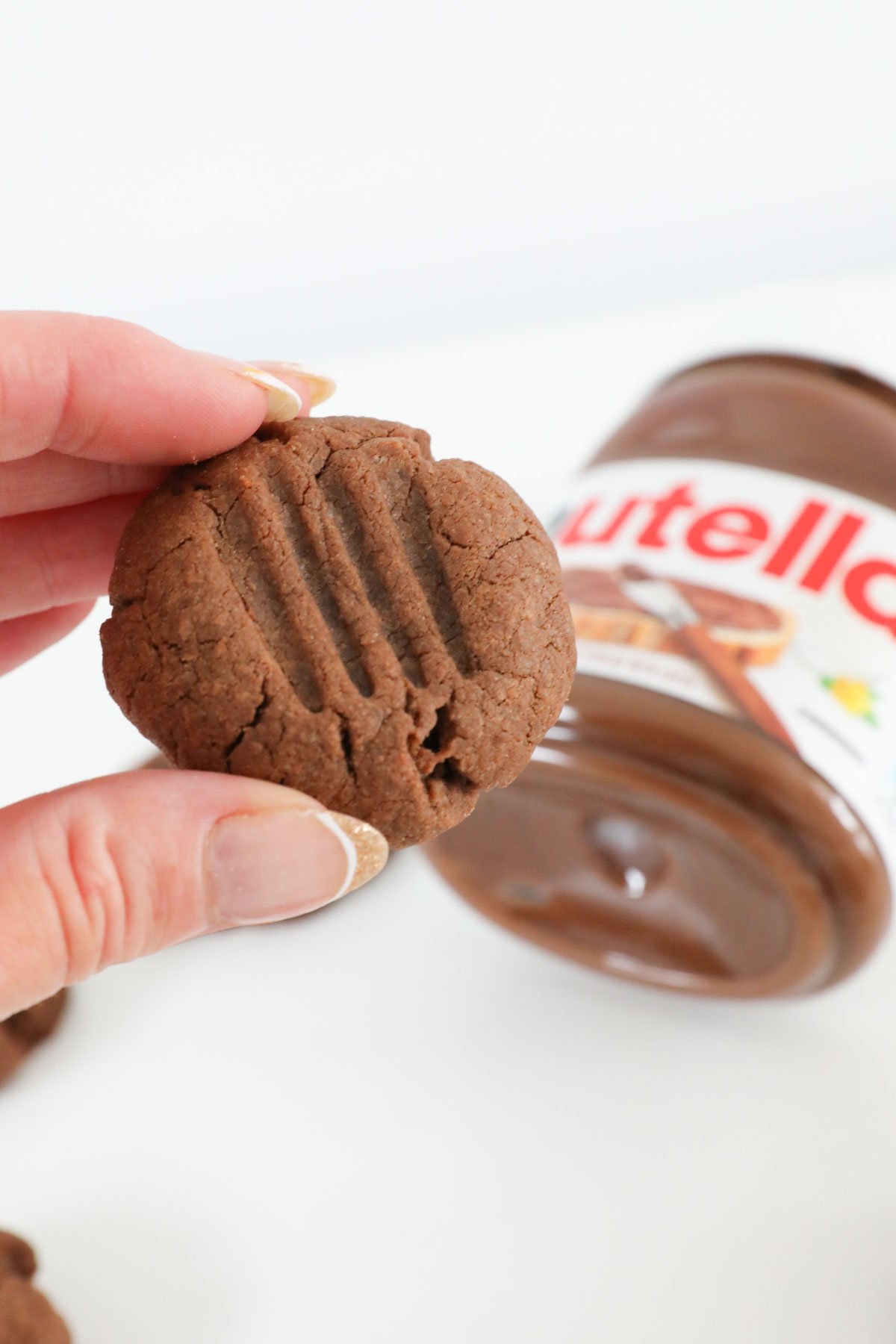 A hand holding up a Nutella cookie, with a jar in the background.