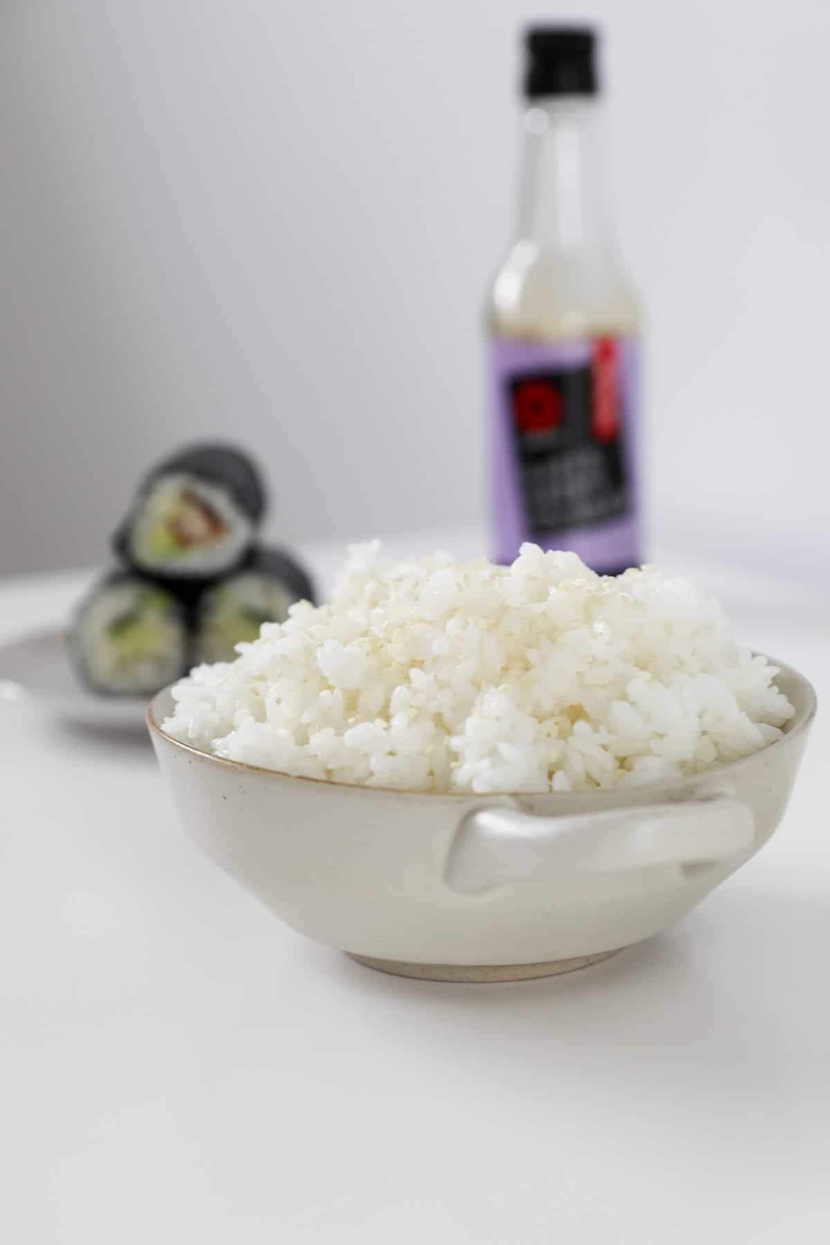 A bowl of rice with soy sauce and sushi rolls in the background.