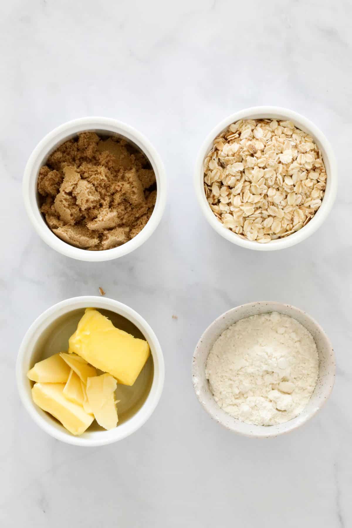 A bowl of brown sugar, a bowl of butter, a bowl of rolled oats, and a bowl of flour.