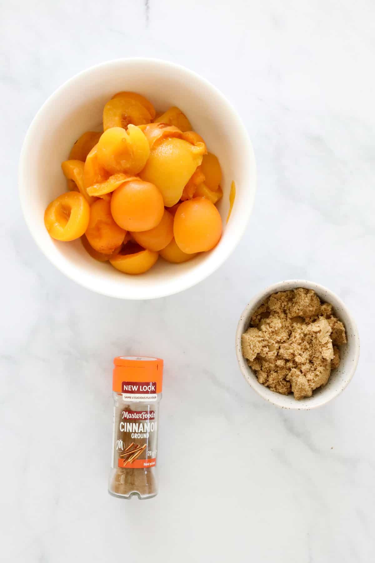 A bowl of apricot halves, small bowl of brown sugar, and a jar of ground cinnamon.