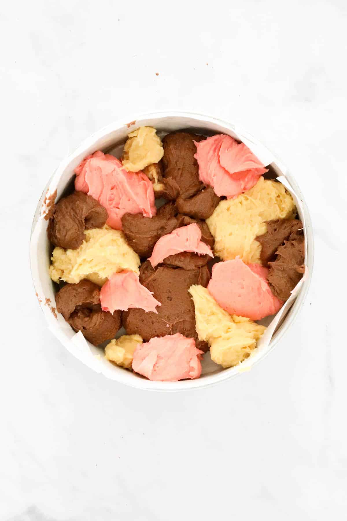 Spoonfuls of vanilla, chocolate and pink cake batter dotted in a round cake tin.
