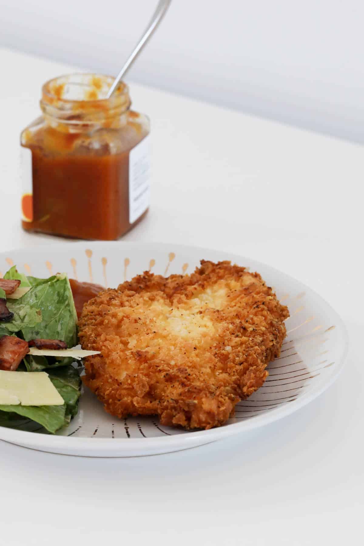 A jar of relish behind a crispy crumbed fillet served with salad.