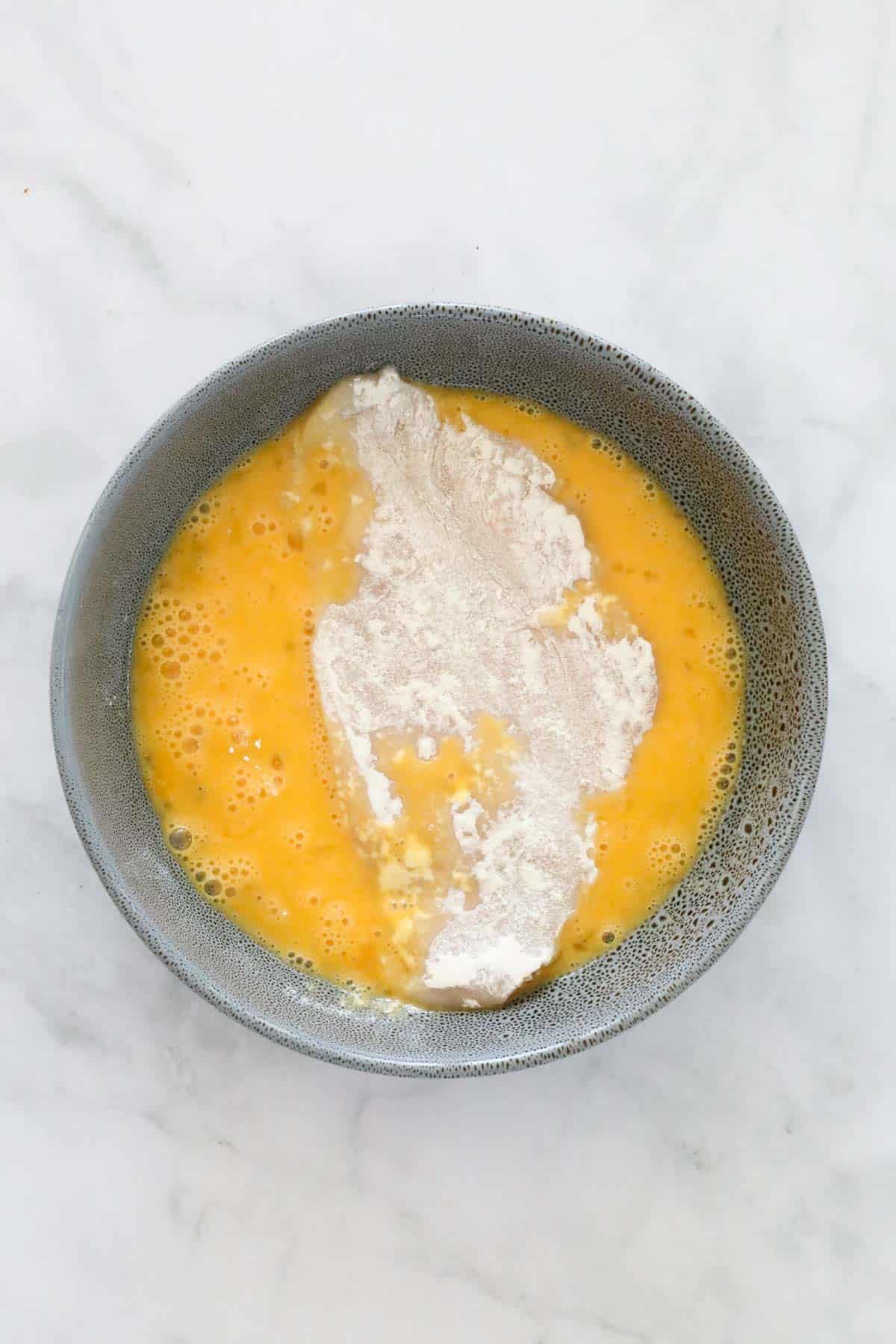 A floured piece of chicken in an eggwash mixture in a bowl.