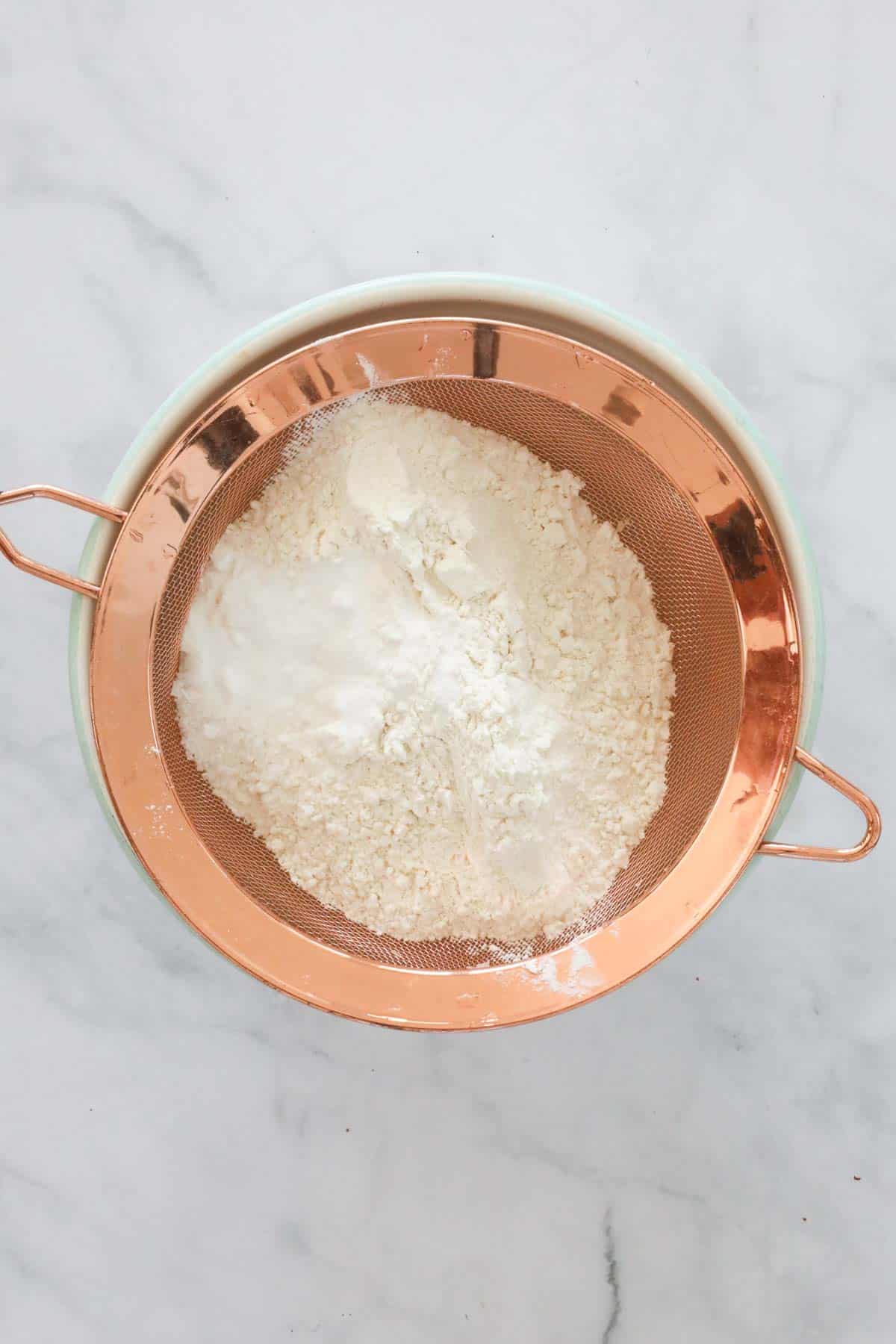Flour and bicarb soda in a copper sieve over a bowl.