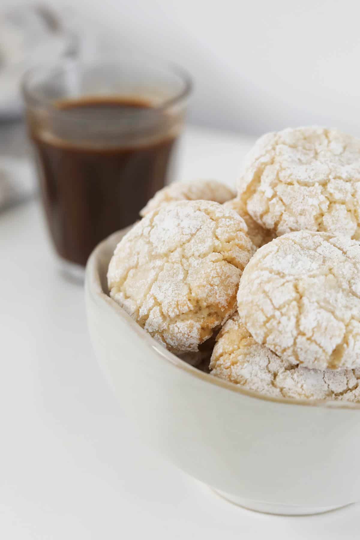 A glass of coffee behind a bowl of sugar coated cookies.