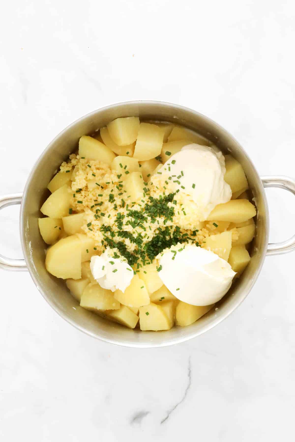 Sour cream, grated cheese and chopped chives added to a saucepan filled with cooked potatoes.