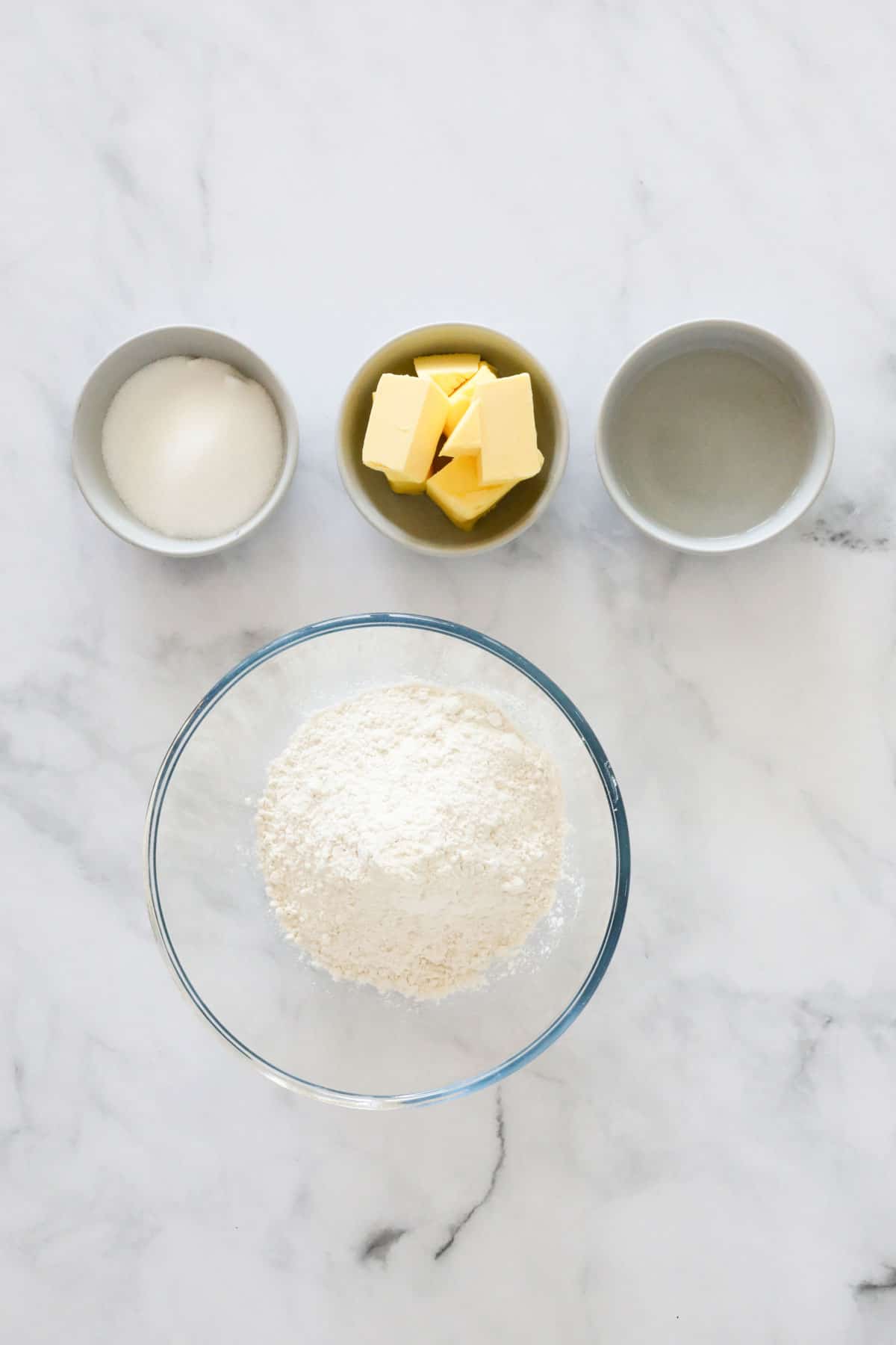 Flour, sugar, butter and water in individual bowls on a bench top.