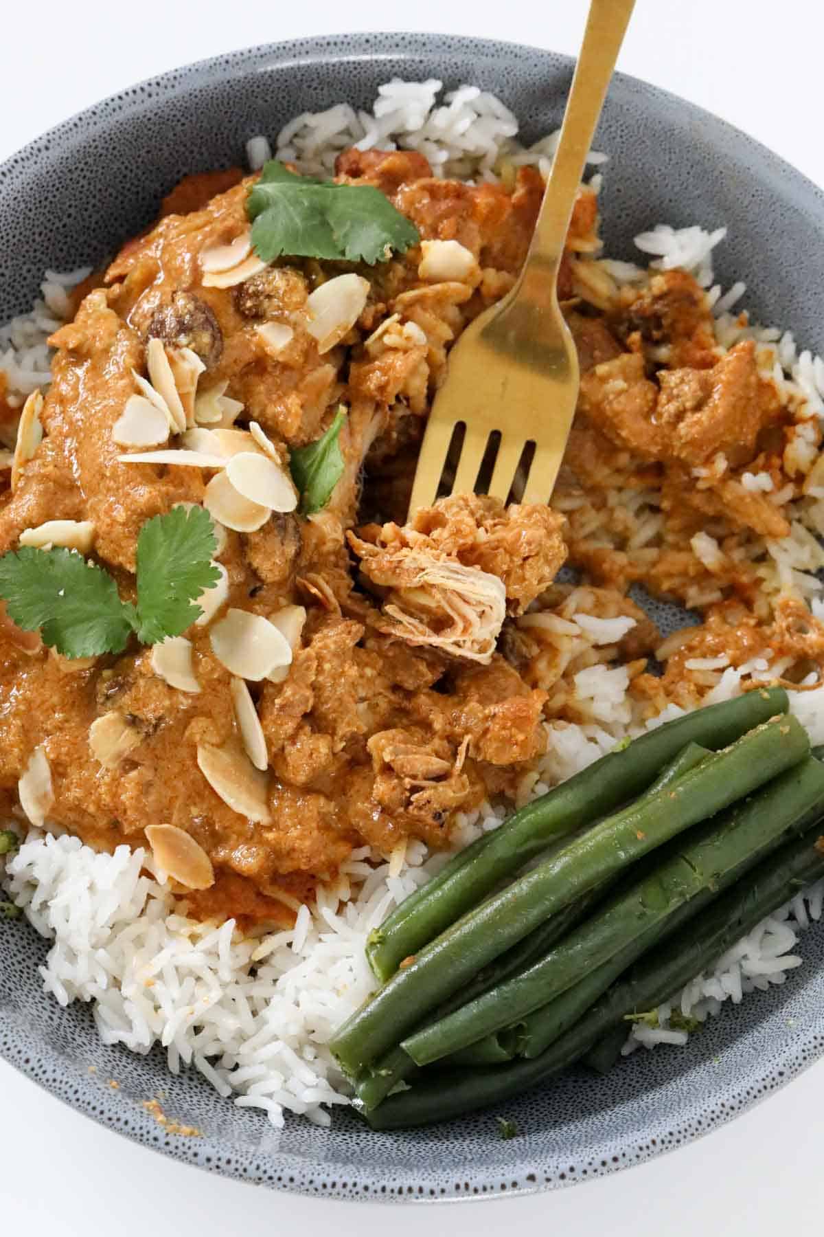 An indian chicken dish served in a bowl with rice and green beans, with a fork lifting a portion of the curry.