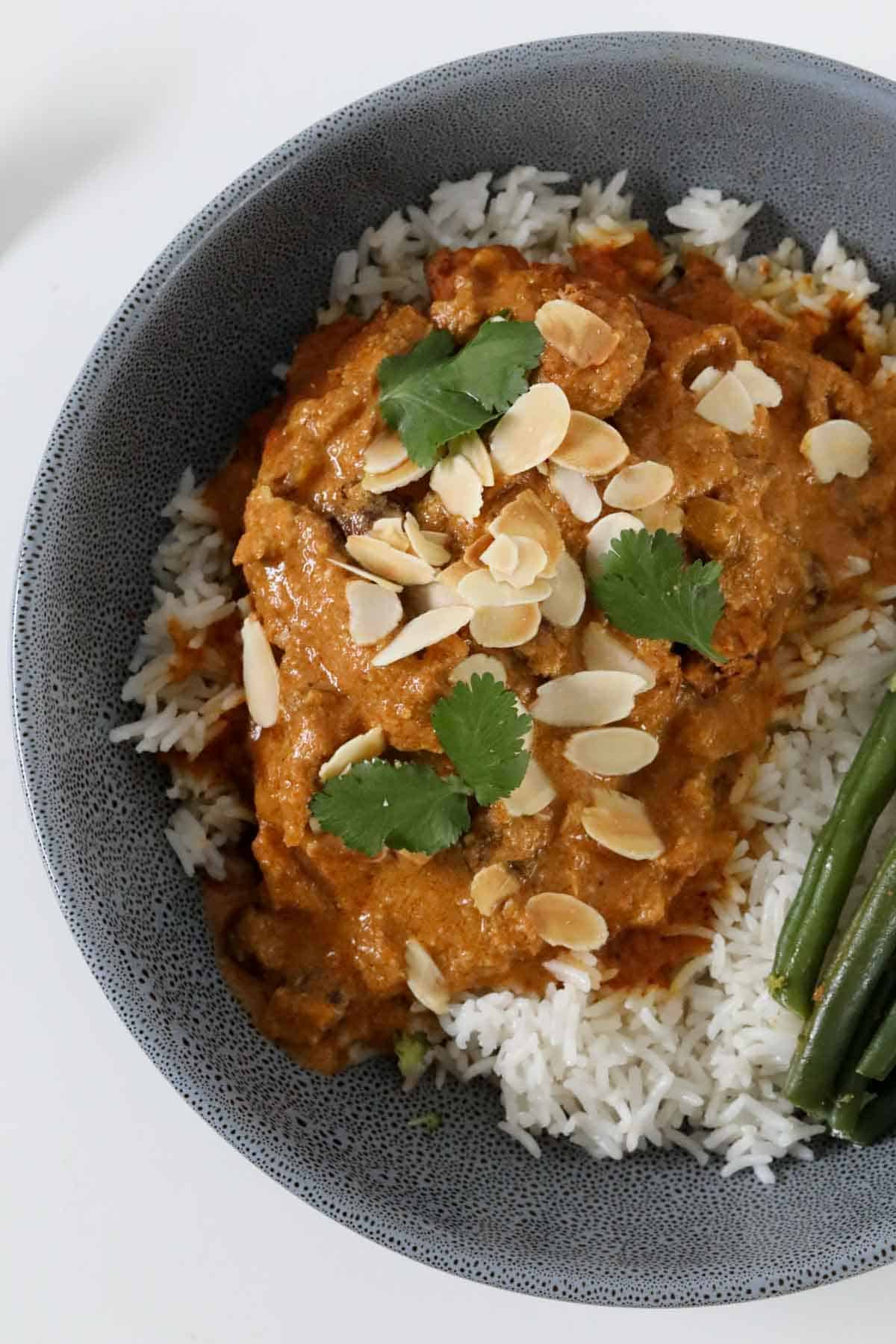 Slow cooker chicken korma served in a grey bowl with rice and green beans, topped with flaked almonds and fresh coriander.