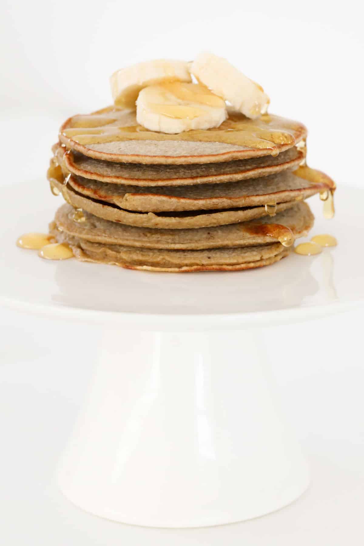 A stack of oat pancakes topped with bananas and honey on a white cake stand.