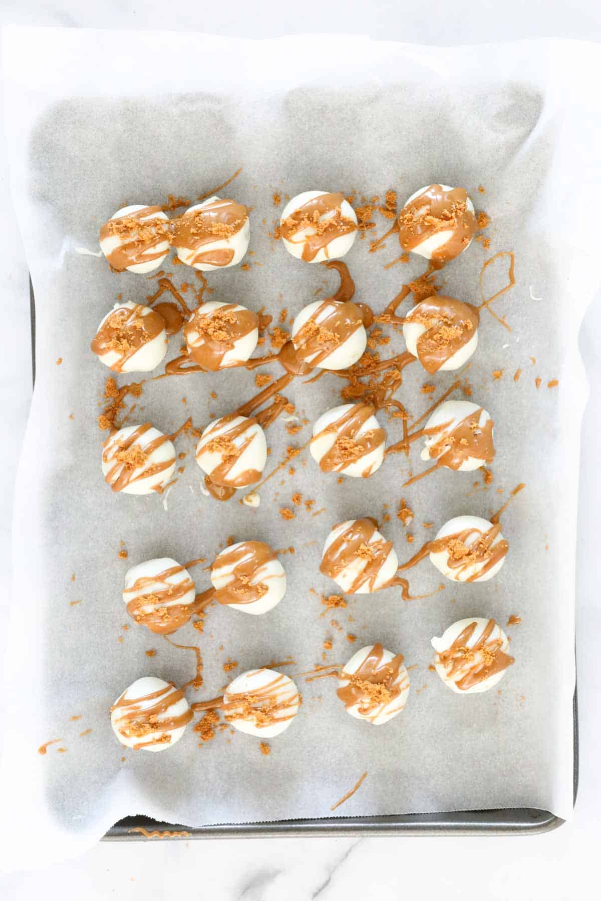 Truffles on the tray dipped in melted white chocolate, drizzled with Biscoff spread and sprinkled with crushed biscuits.