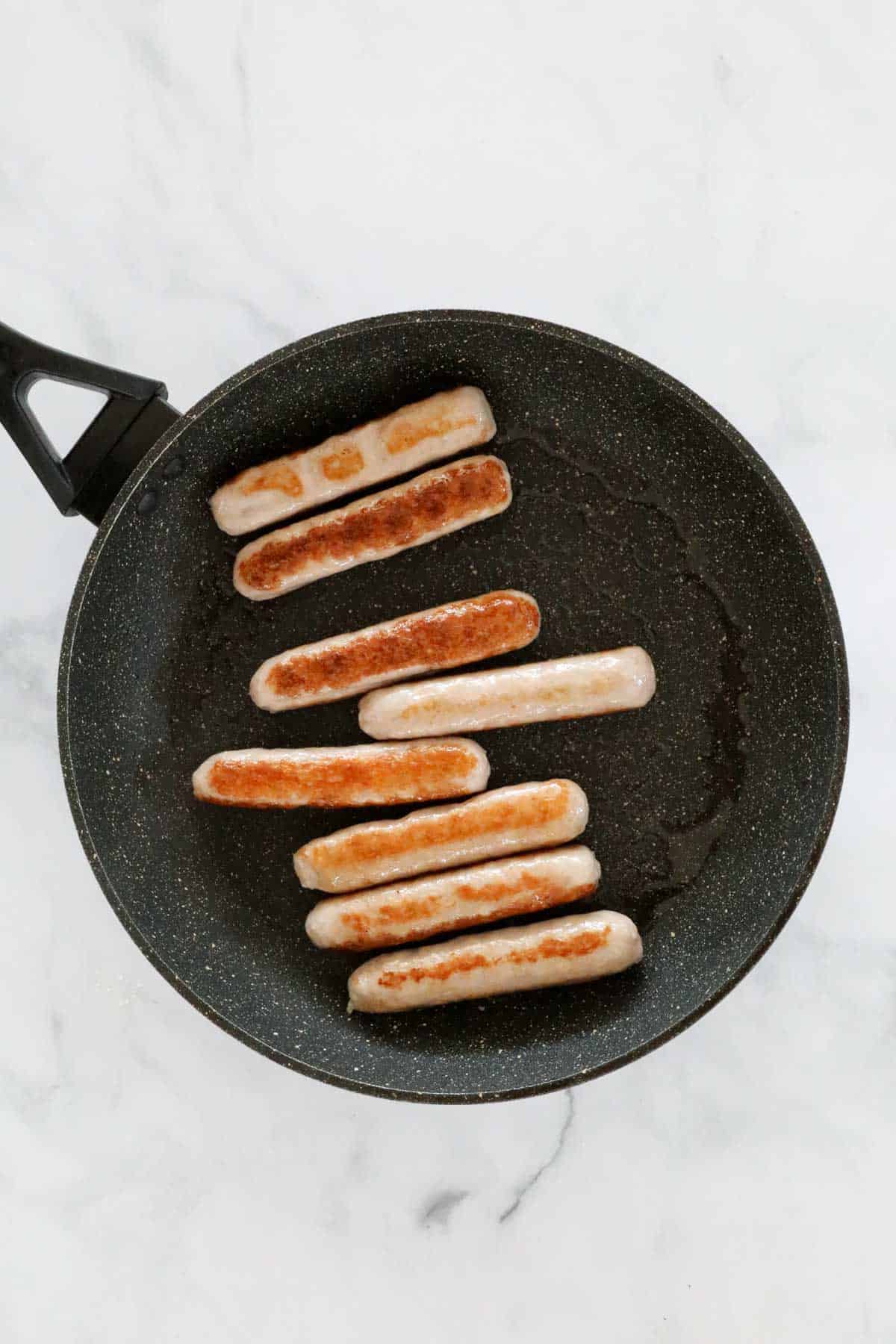 Thin sausages browning in a frying pan.