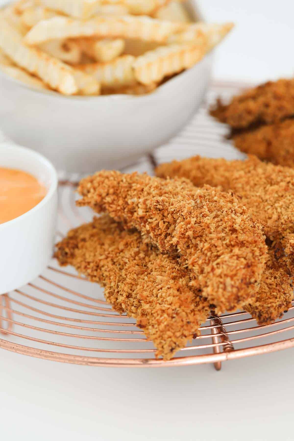 A pile of baked chicken tenderloins on a copper tray with a dipping sauce beside them.