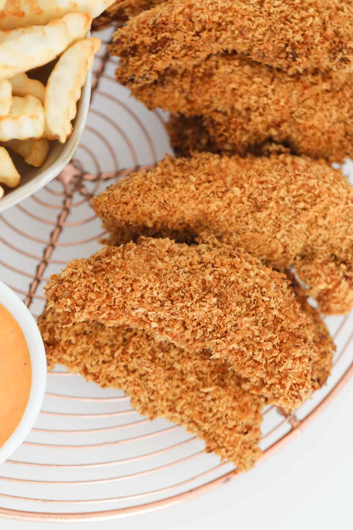 Crispy crunchy baked strips on chicken on a wire rack with a small bowl of dipping sauce.