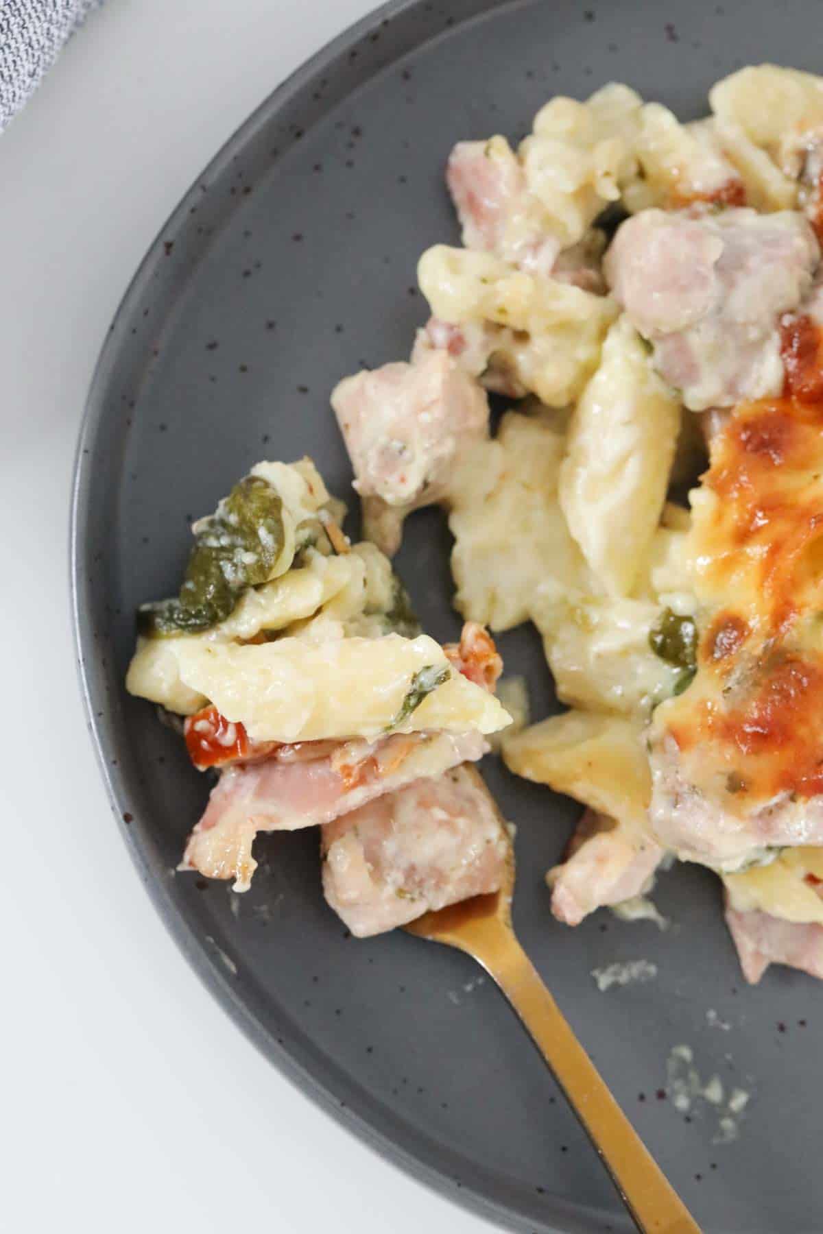 A gold fork on a plate with a serve of creamy chicken and bacon pasta bake.