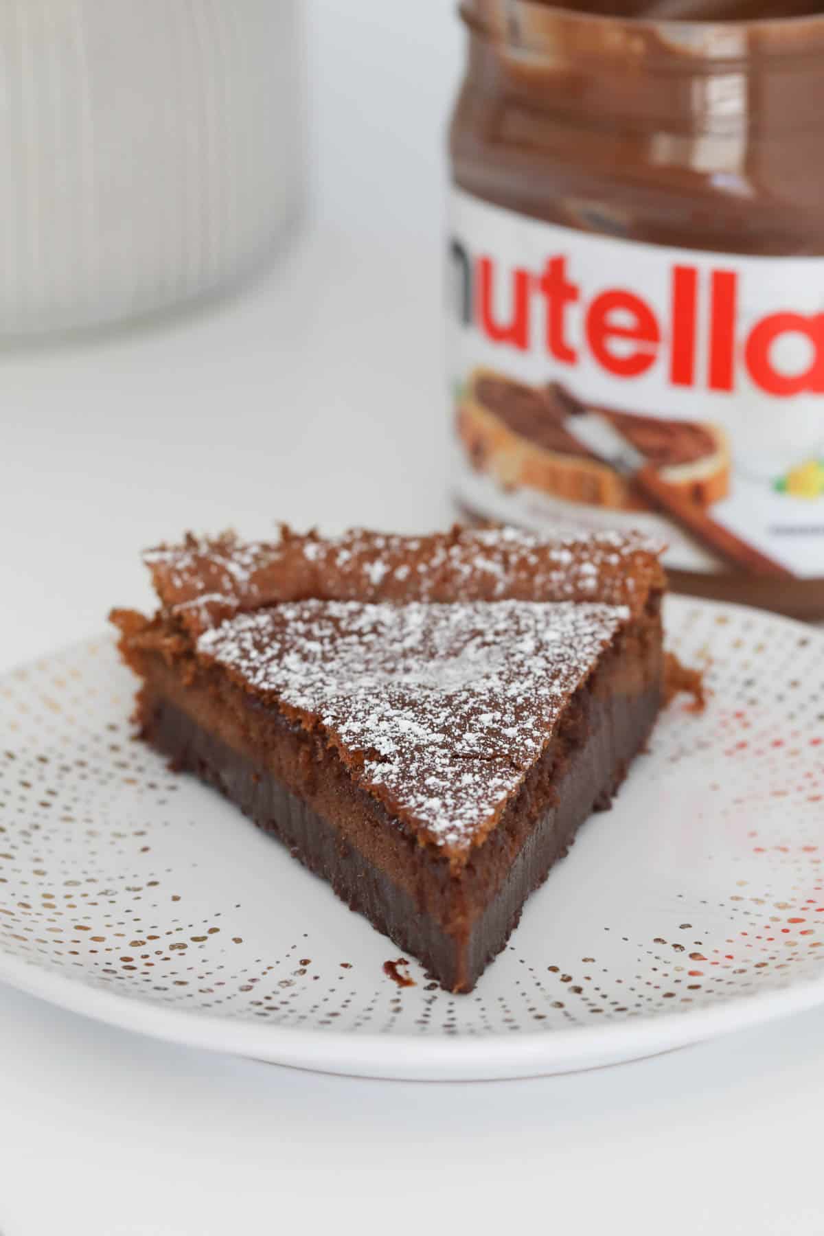 A slice of baked fudgy cake on a plate with a jar of Nutella in the background.