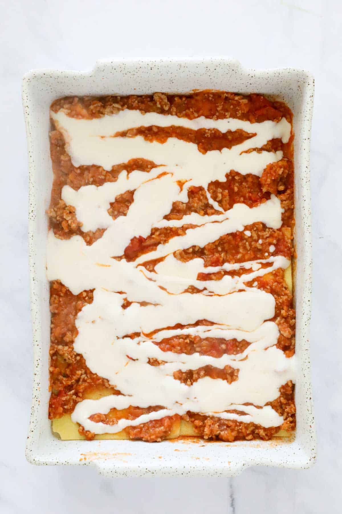 Bechamel sauce drizzled over bolognese sauce in a baking dish.
