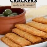 A pile of crunchy cheesy crackers with a bowl of green olives behind them.