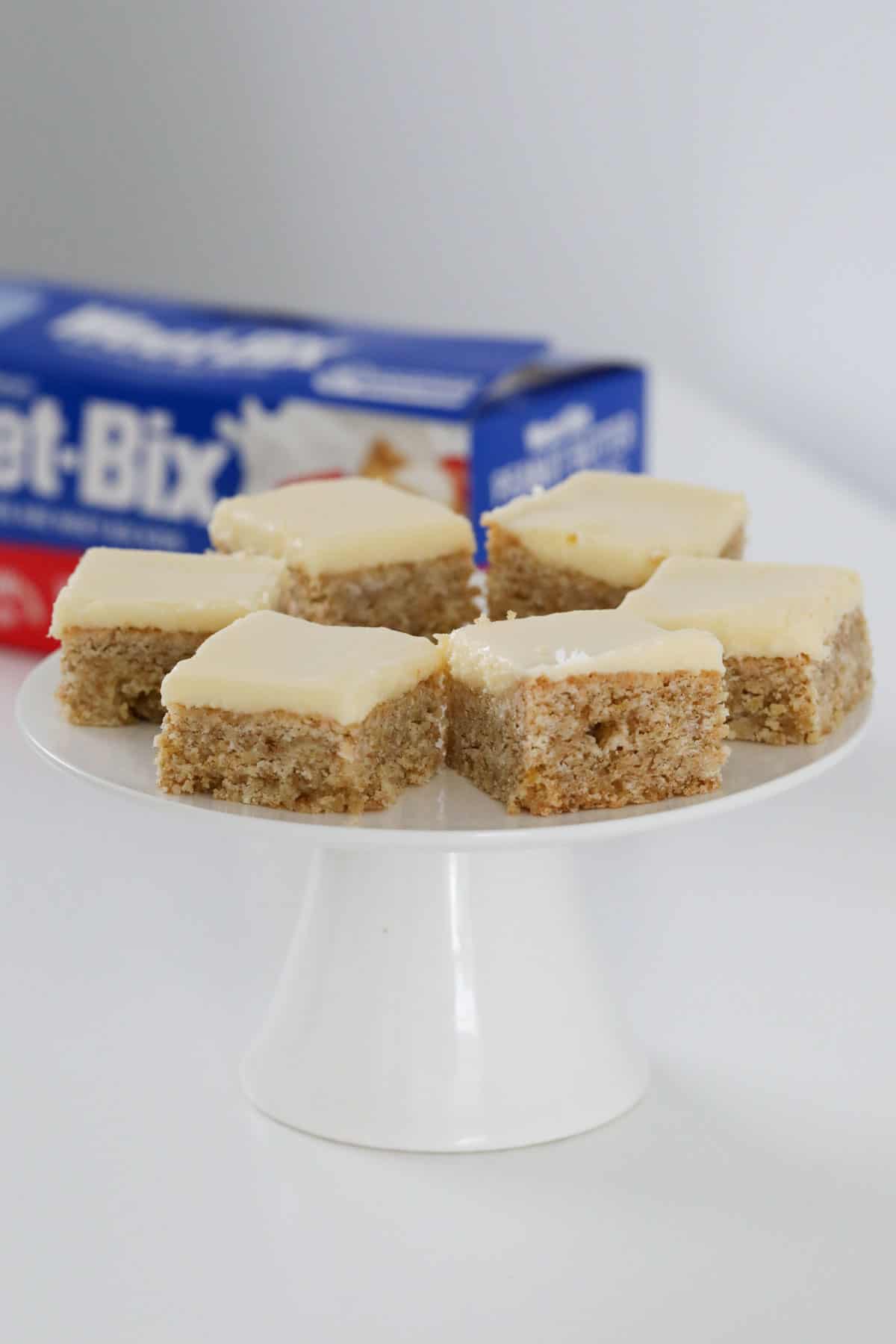 A cake stand with squares of lemon slice on it, in front of a box of Weet-Bix.