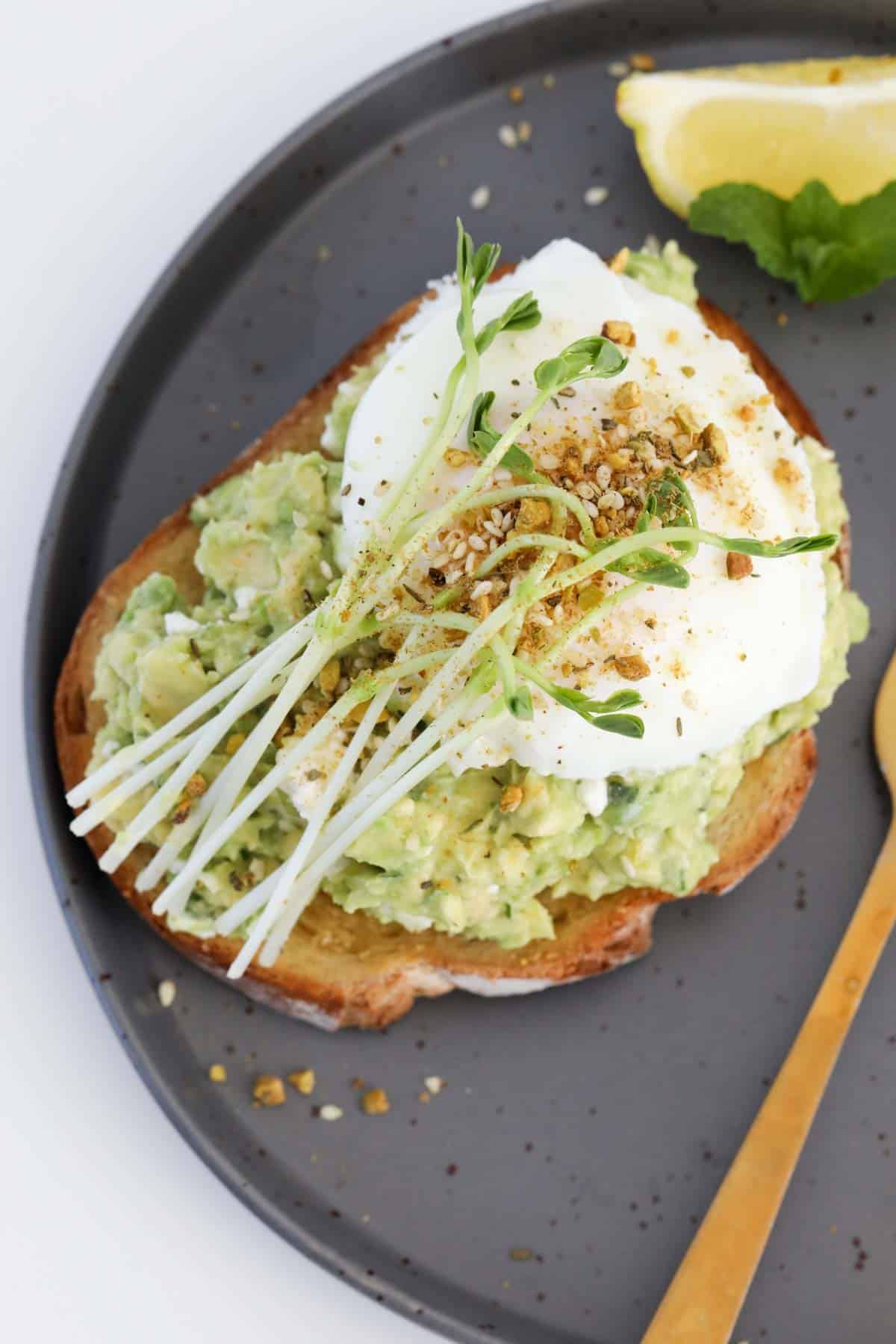 A slice of smashed avocado on toast, served on a black plate topped with a poached egg.