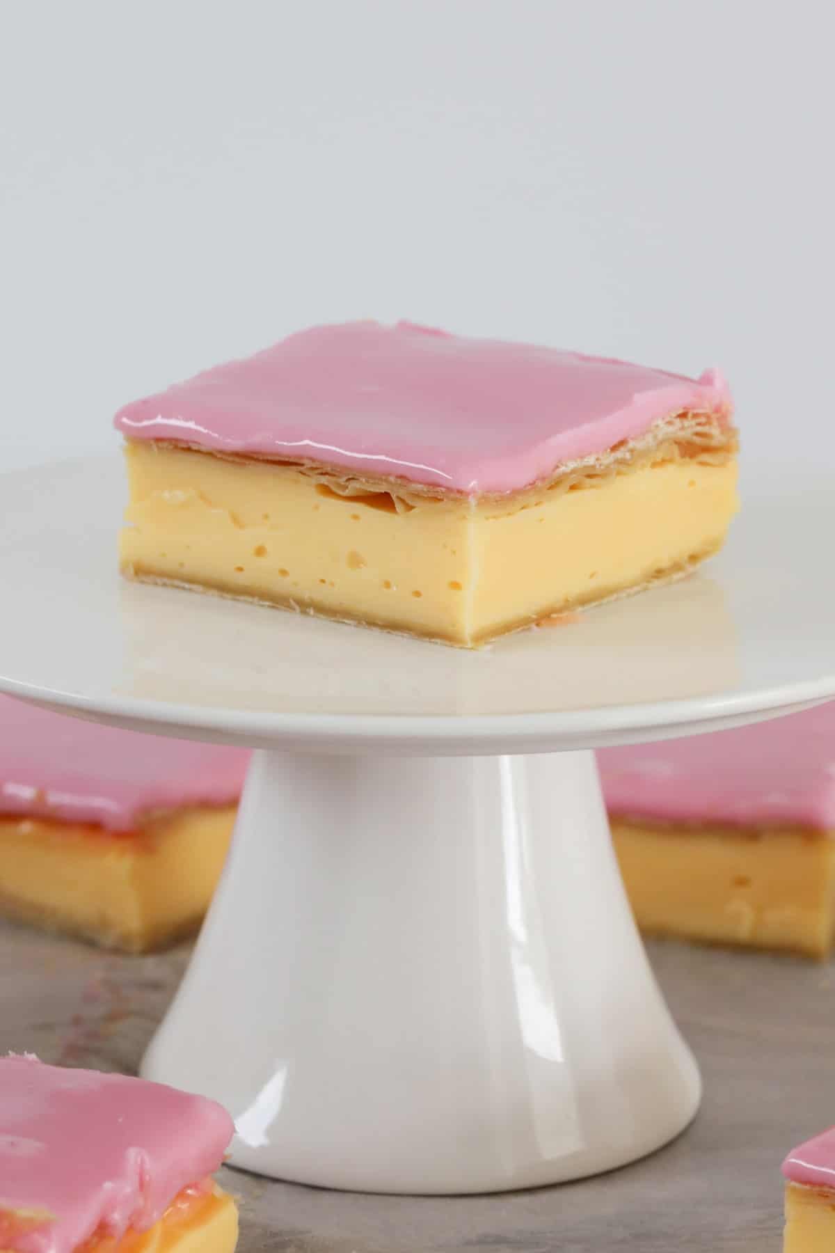 A pink glazed vanilla custard square on a white cake stand.