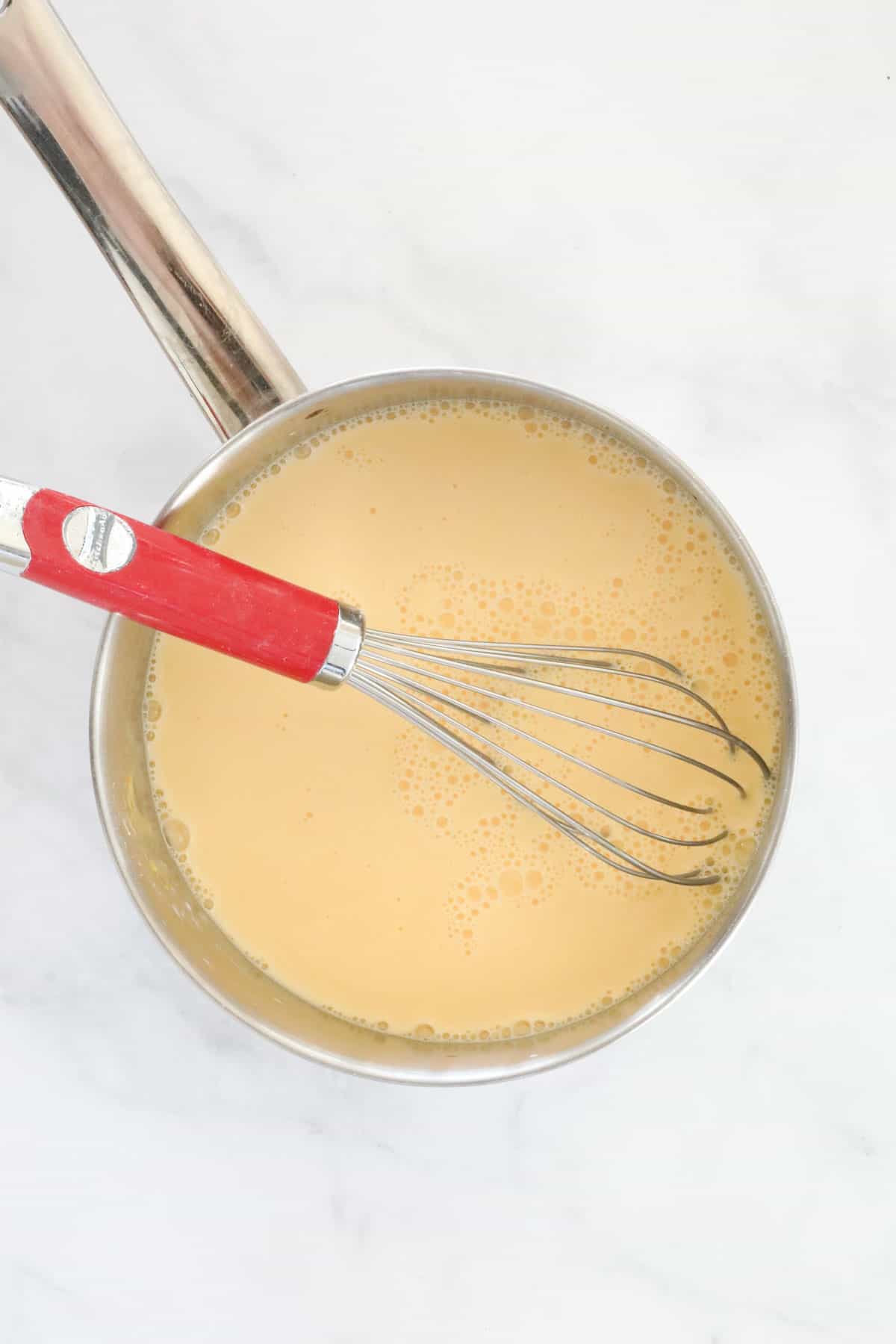 A red handled whisk in a pot of custard powder, sugar and milk.