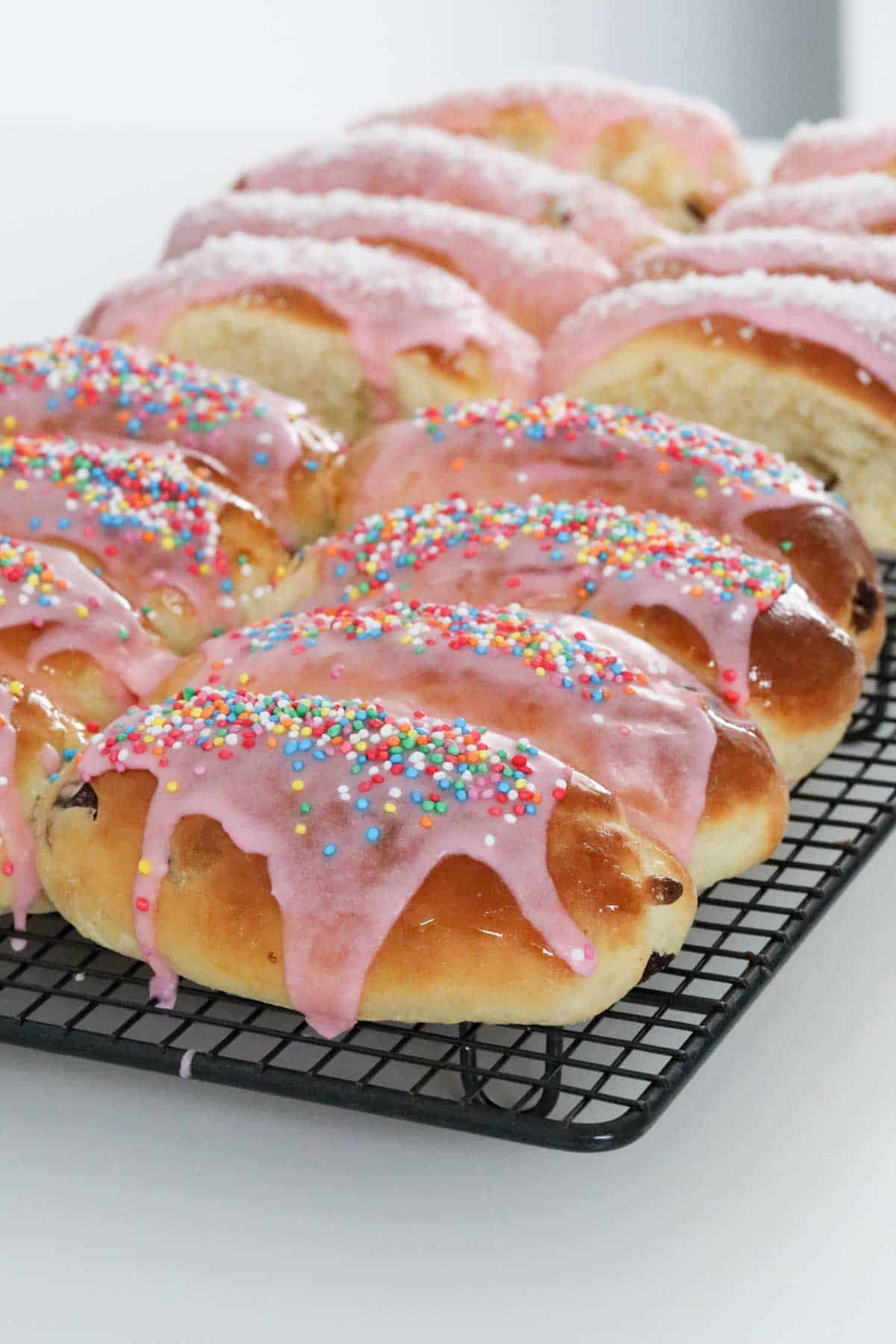 baked finger buns on a cooling rack iced with pink icing and some sprinkled with coconut, others sprinkled with 100s and 1000s.