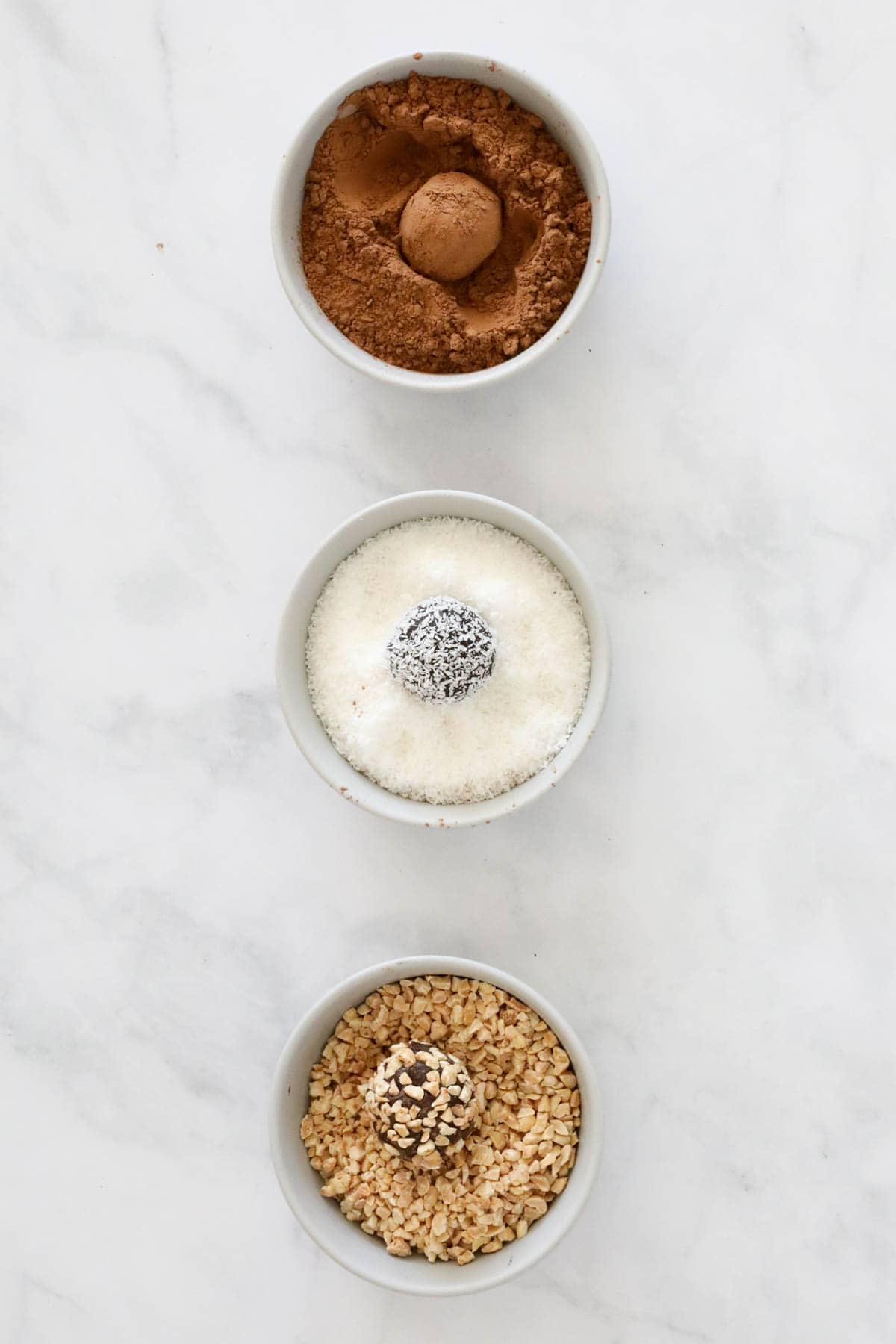 Three bowls of truffle coatings: cocoa powder, coconut and chopped peanuts, with a ganache truffle in each one.
