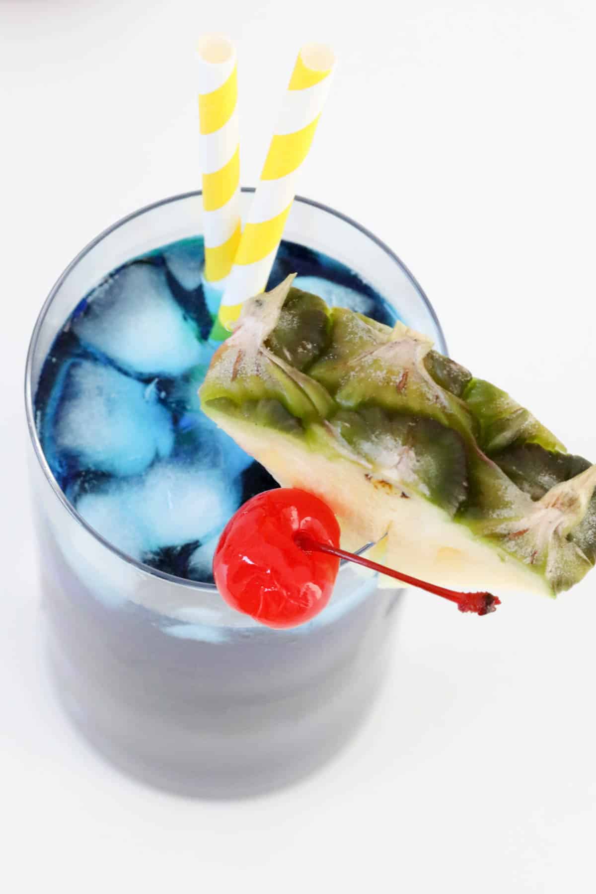 An overhead shot of a blue cocktail in a glass with a pineapple and cherry.