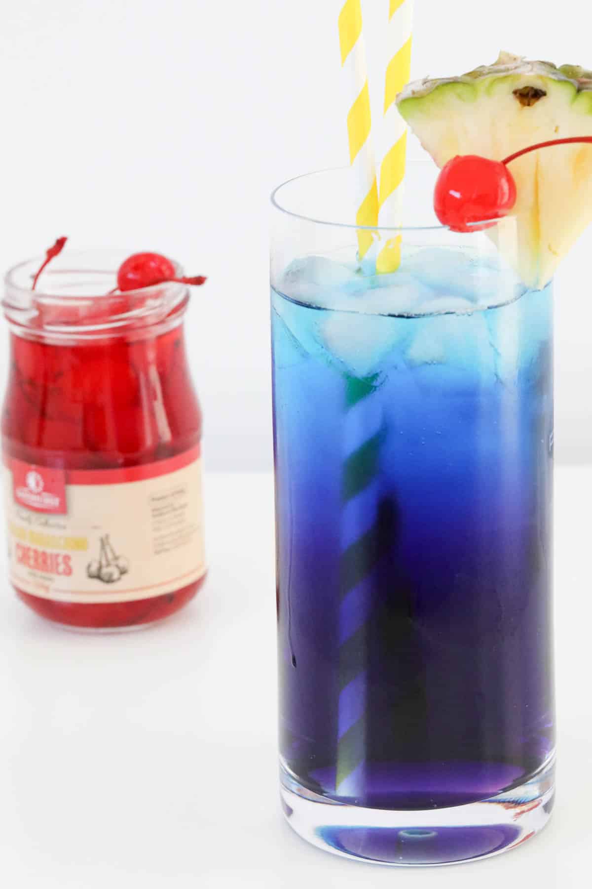 A jar of maraschino cherries behind a blue tingle cocktail.