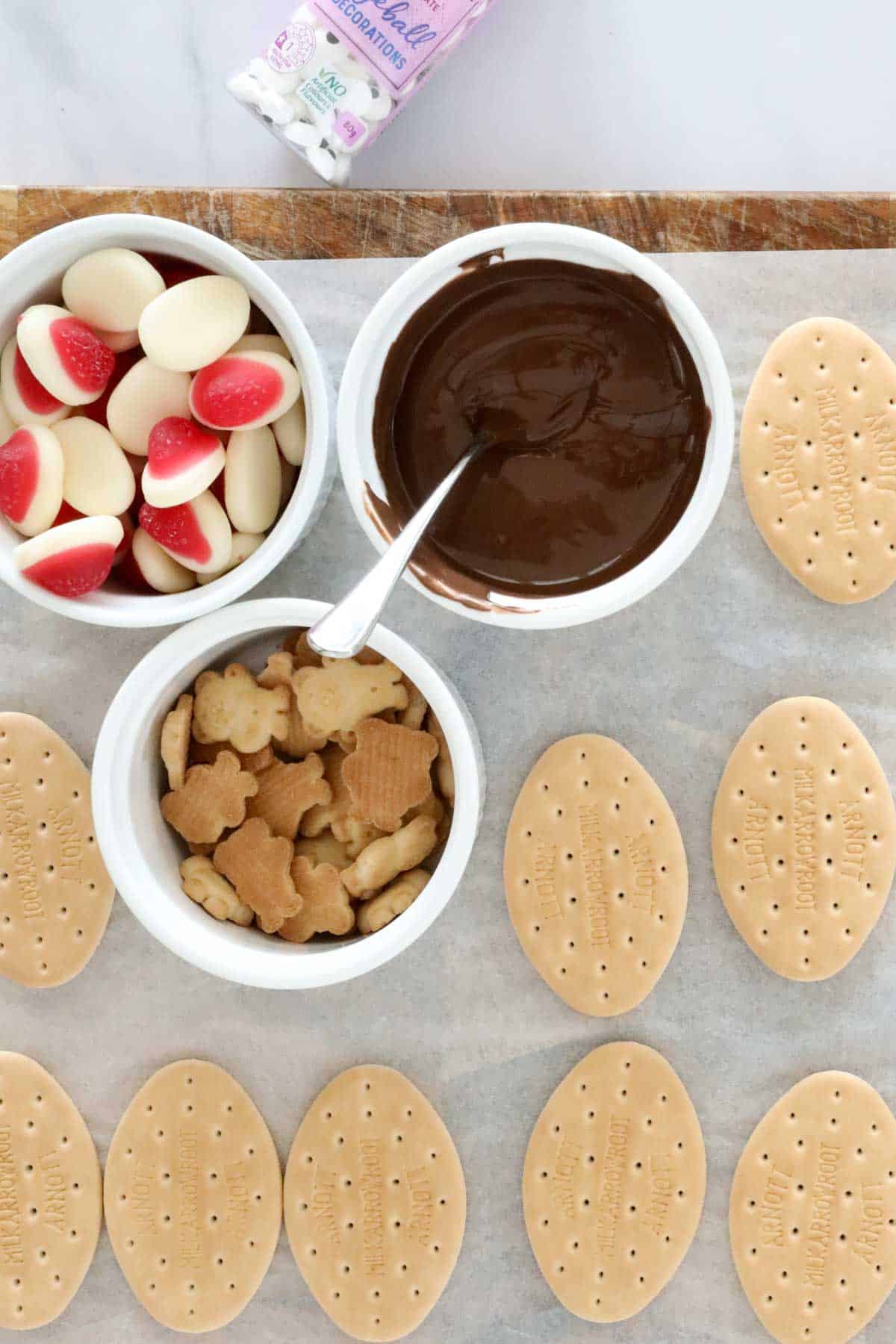 Milk arrowroot cookies placed on a lined tray, with bowls of melted chocolate, jelly sweets and Tiny Teddys.