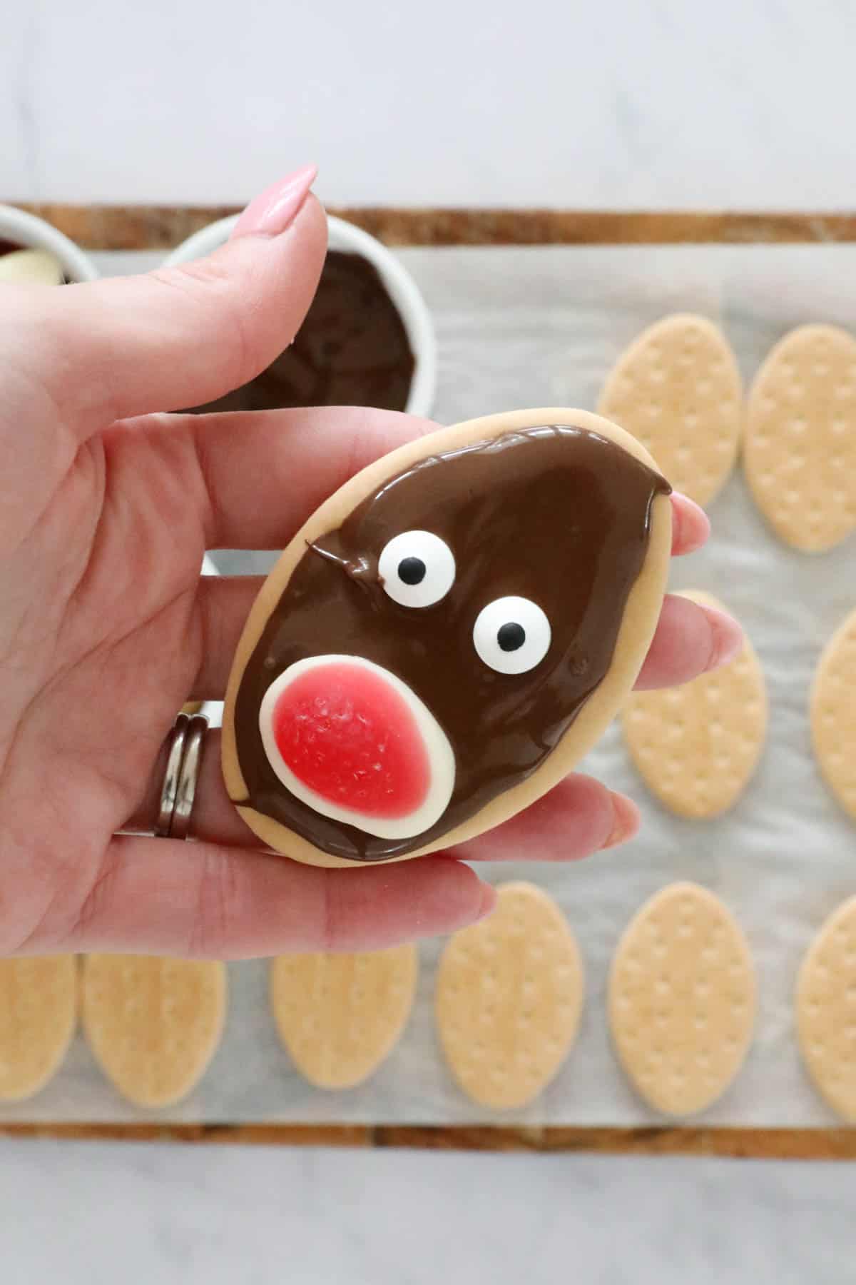 A hand holding a biscuit covered in melted chocolate with two candy eyes and a jelly sweet red nose.
